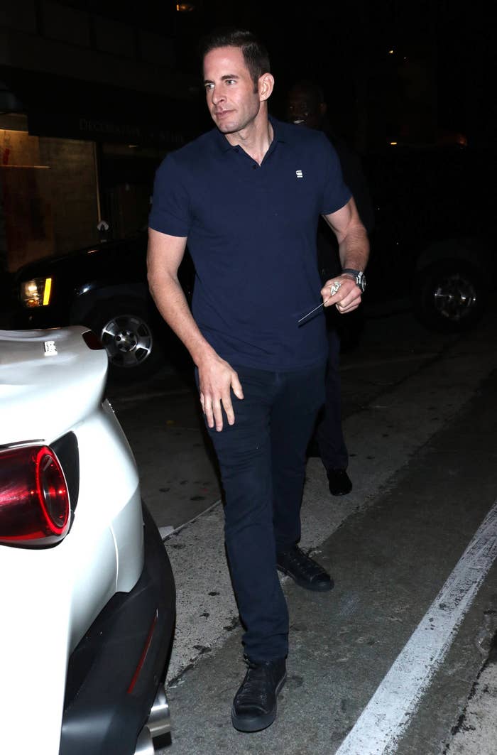 Tarek El Moussa is seen wearing a dark shirt and pants, walking by a car at night