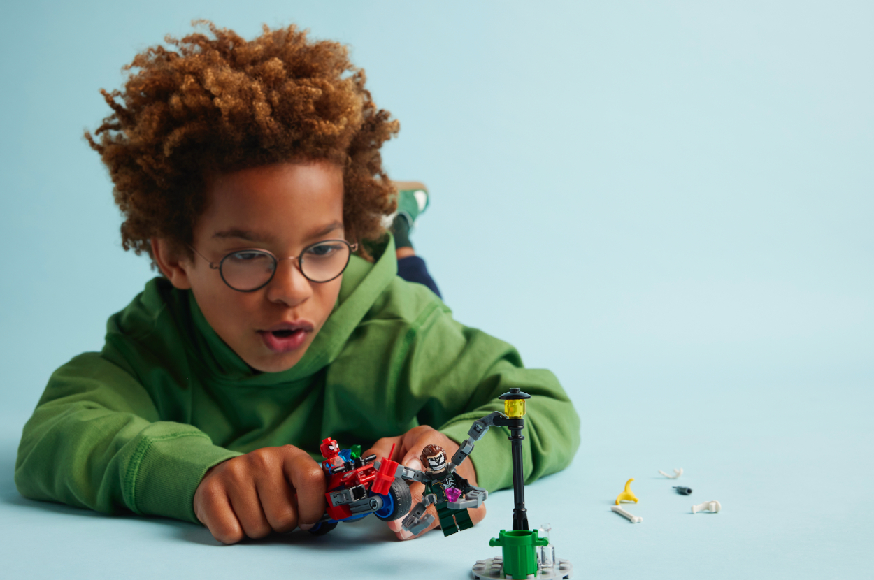 Young boy with curly hair and glasses wearing a green hoodie plays with Lego figures on the floor