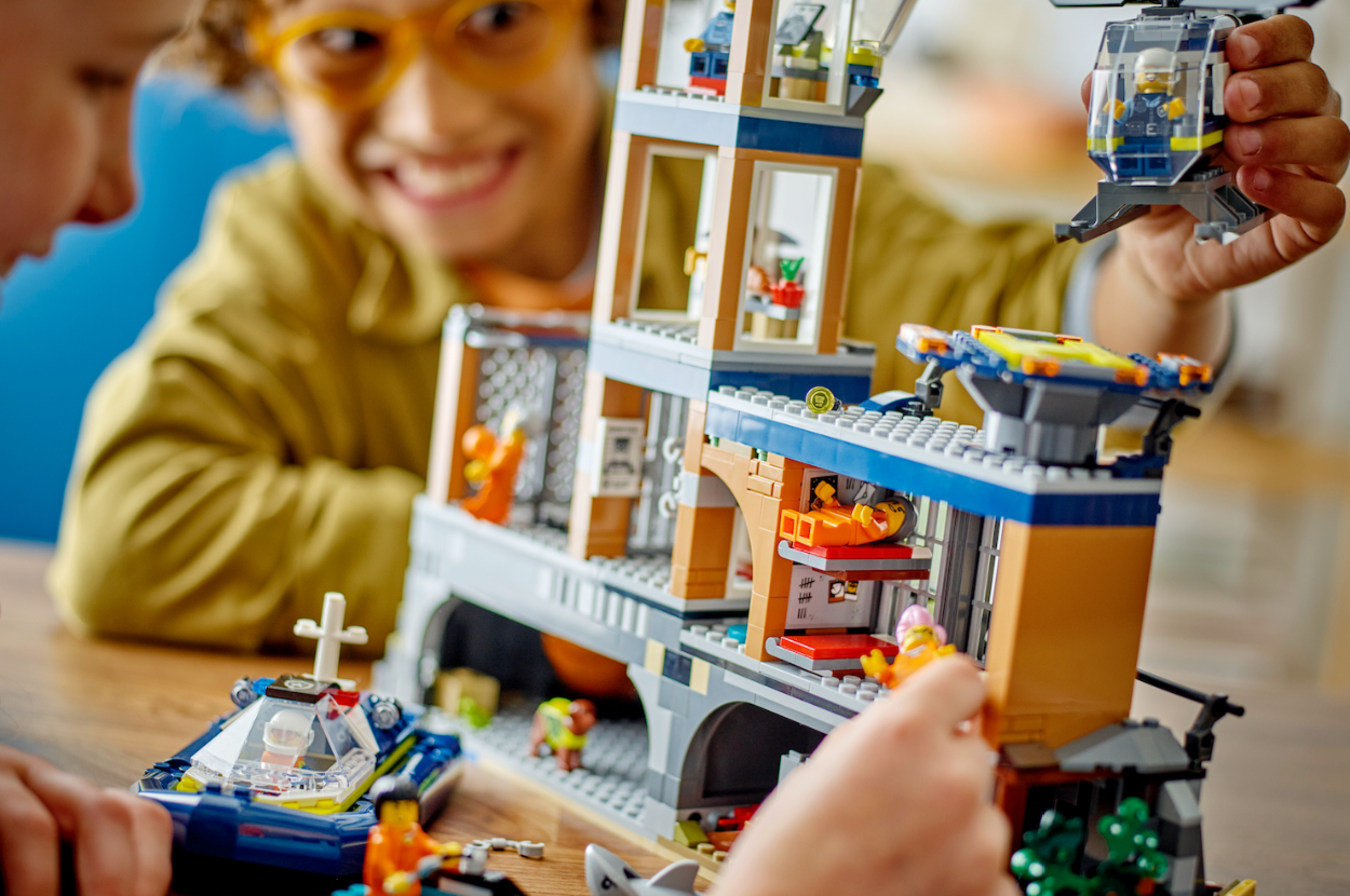 Children playing with a LEGO set, assembling a building, police boat, and helicopter. Faces of children smiling are visible