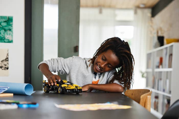 A young child with long hair smiles while playing with a toy car on a table in a bright, cozy room
