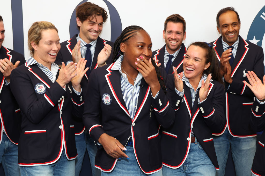 Coco Gauff, clad in a navy blazer with a red trim, laughs and is surrounded by people applauding in similar attire at a celebrity event