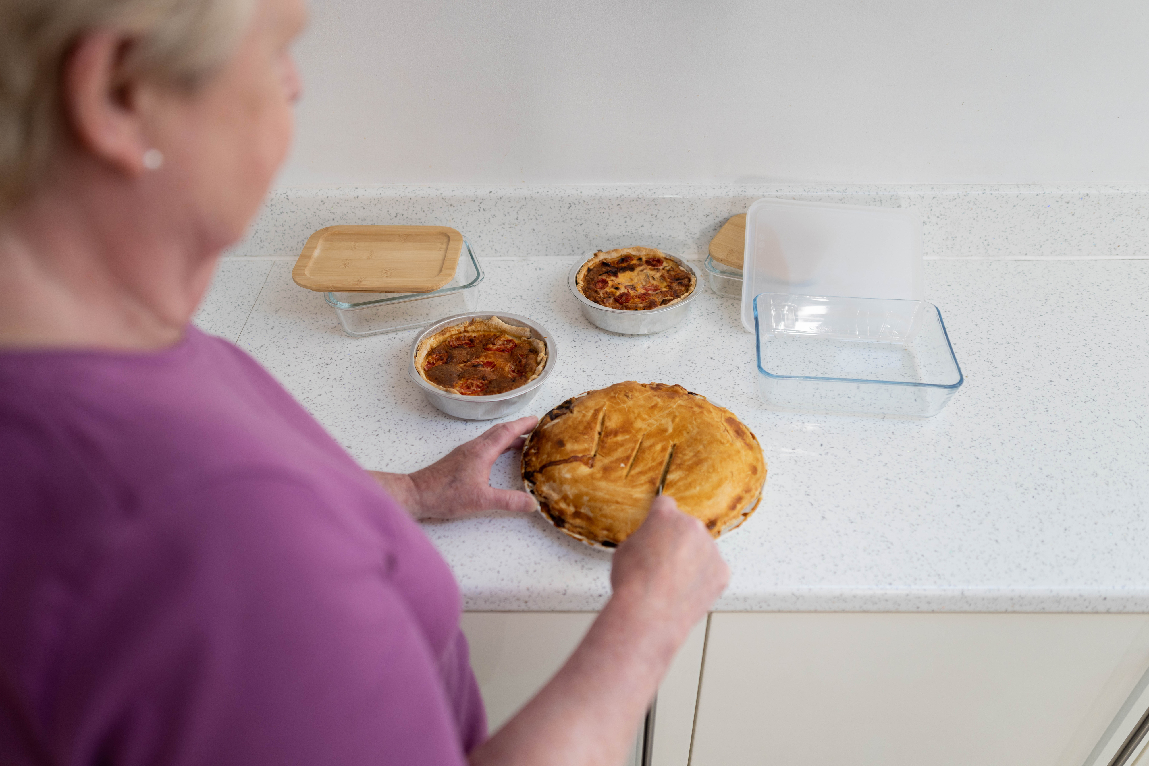 A woman is slicing a freshly baked pie on a kitchen counter with two smaller pies and a glass baking dish nearby
