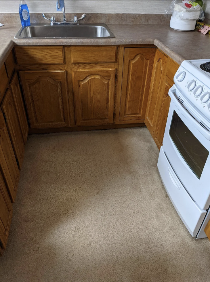 Small kitchen with wooden cabinets, a stainless steel sink, stove, and light-colored carpet flooring. Detergent bottle and paper towels are on the counter