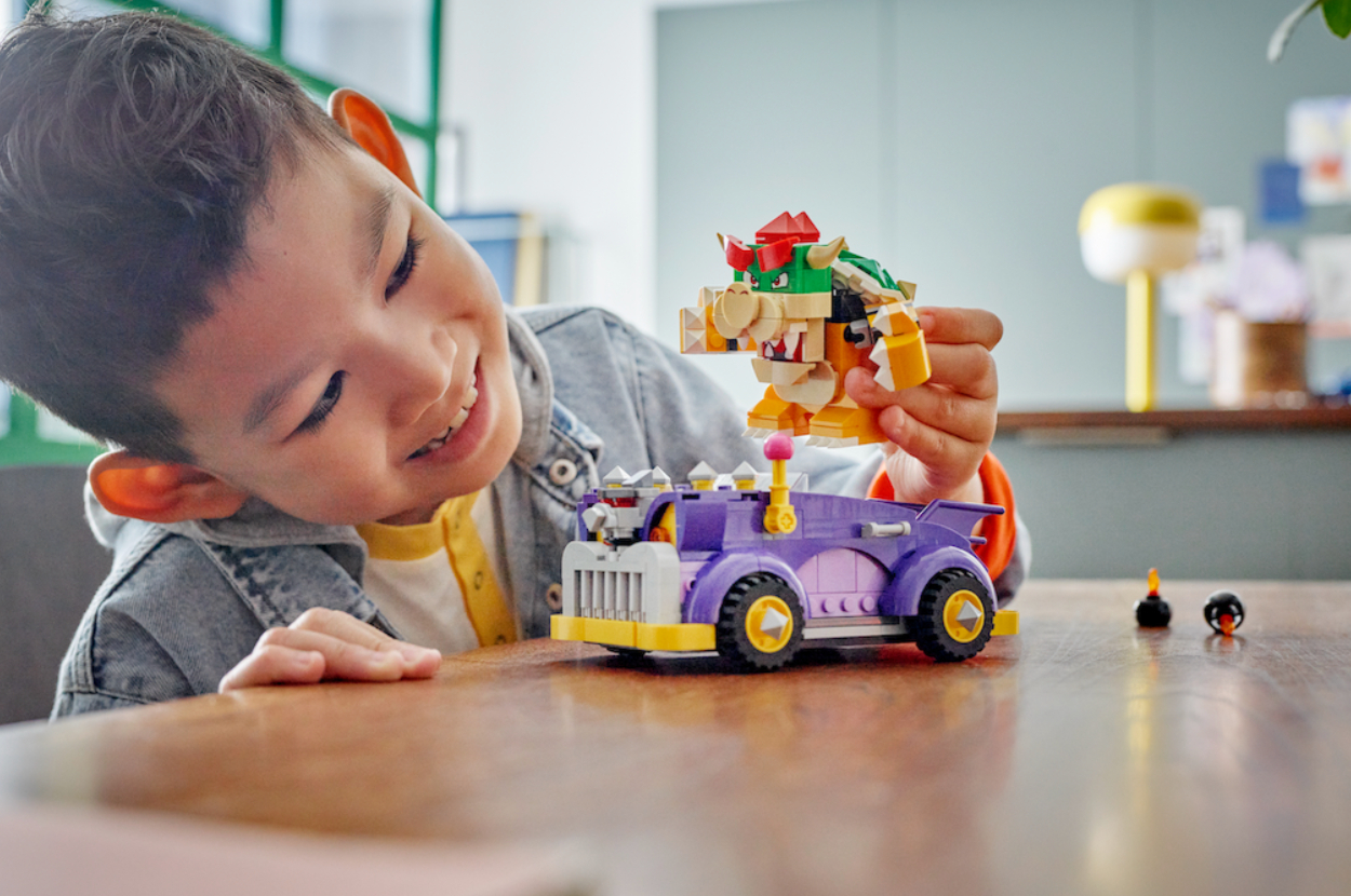 A child is smiling and playing with a LEGO Super Mario Bowser’s Castle Boss Battle set on a wooden table
