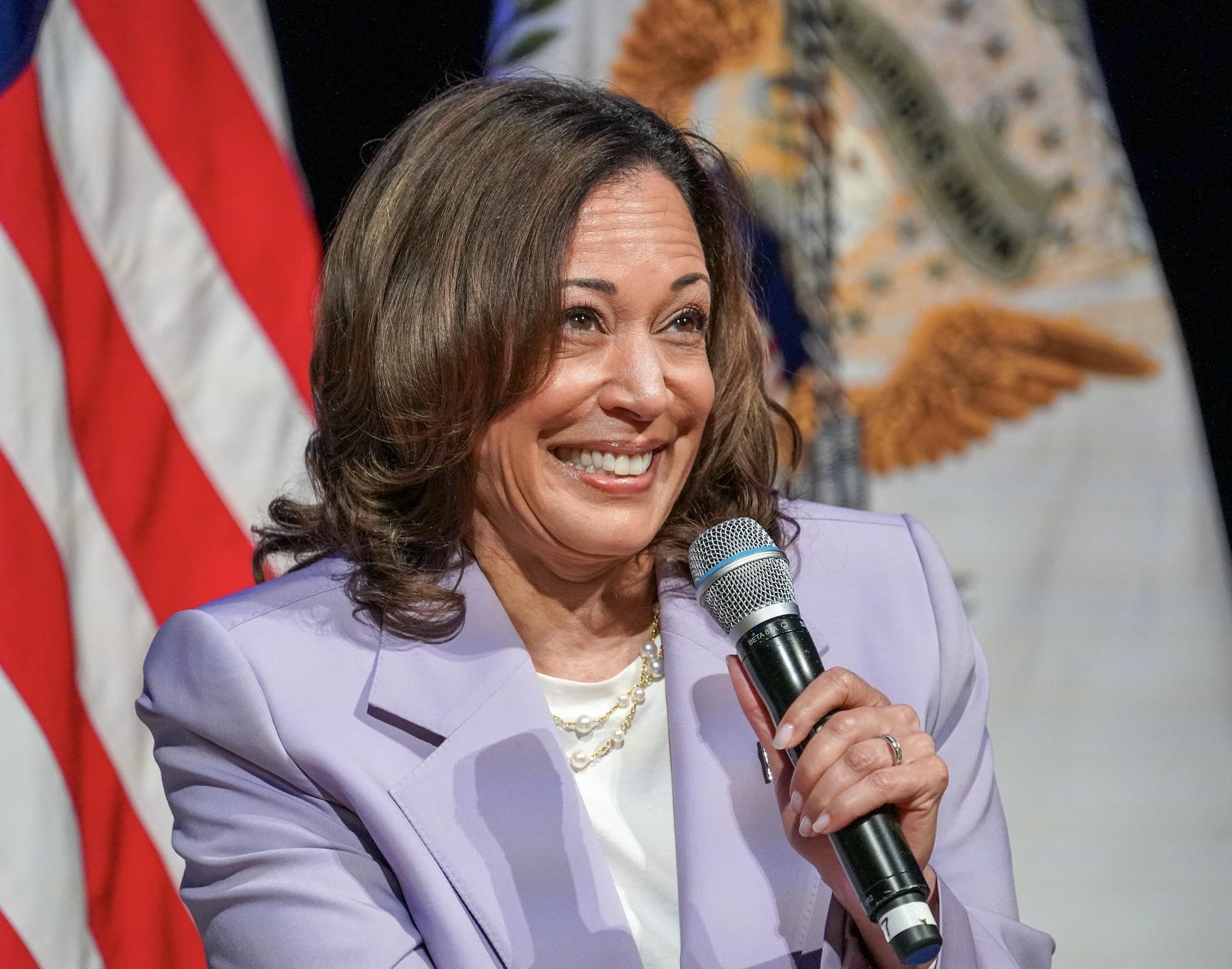 Kamala Harris smiling and holding a microphone, wearing a light-colored blazer and a pearl necklace, with U.S. flags in the background
