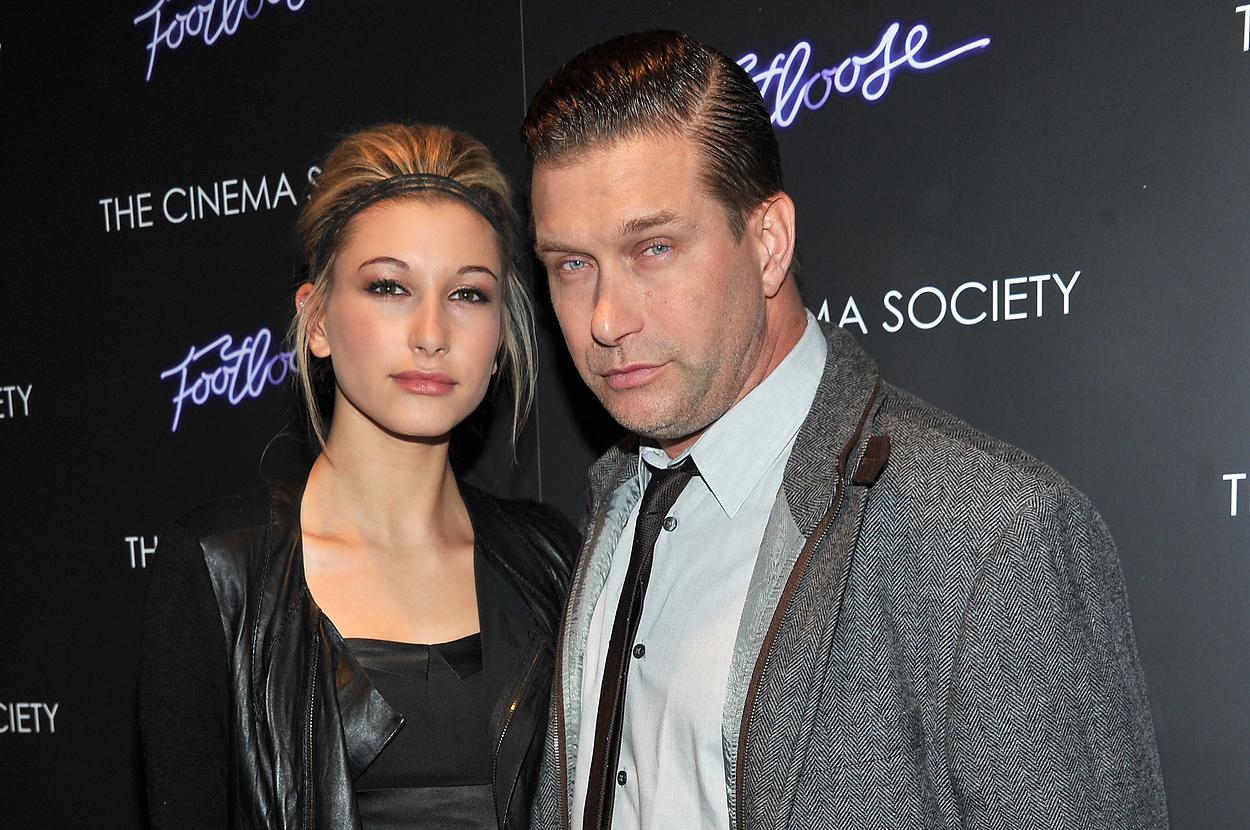 Hailey Bieber in a ruffled black dress and leather jacket, posing with Stephen Baldwin, who is wearing a gray jacket over a white shirt and tie, at a Footloose event
