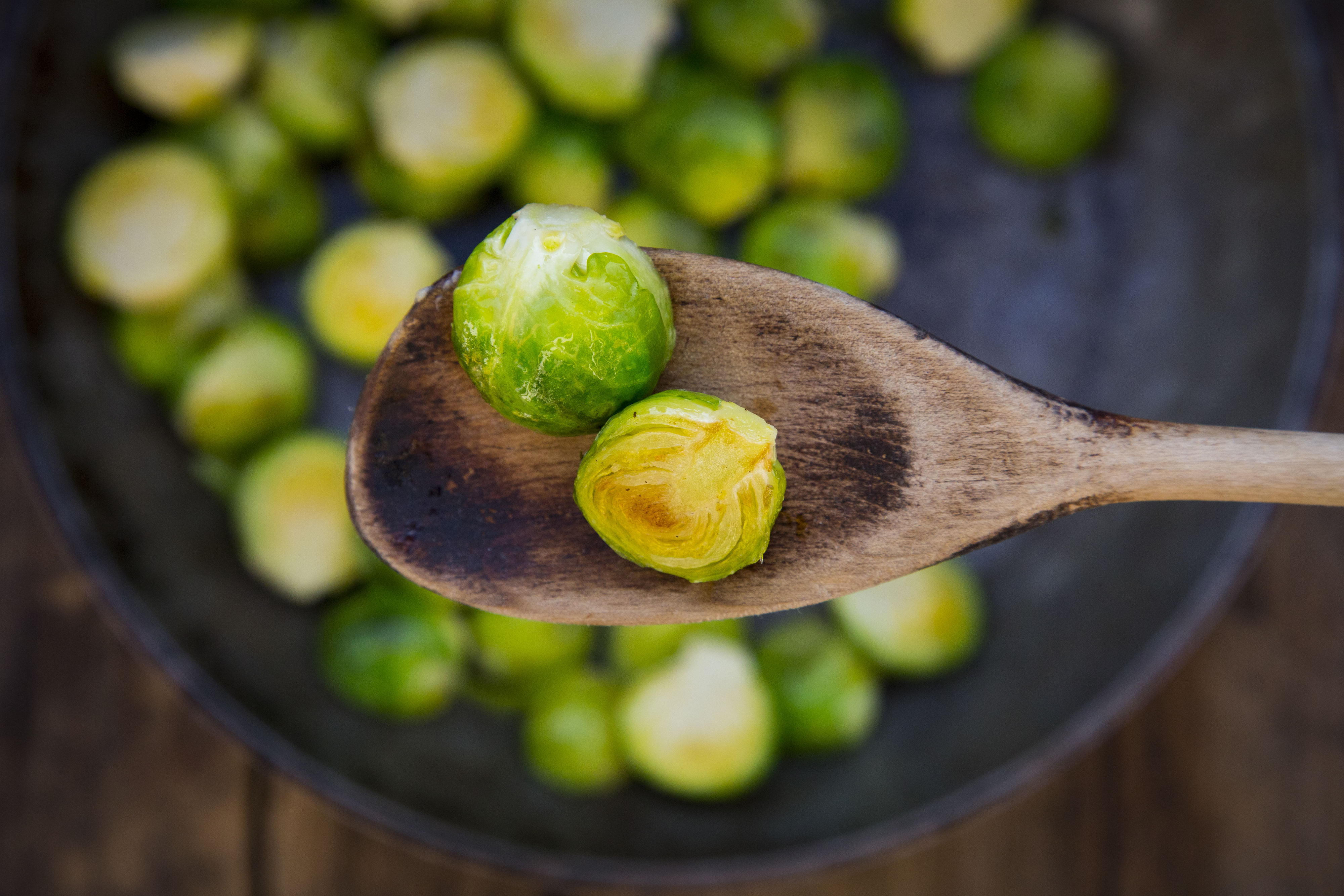 A wooden spoon holds two cooked brussels sprouts over a pan filled with more brussels sprouts