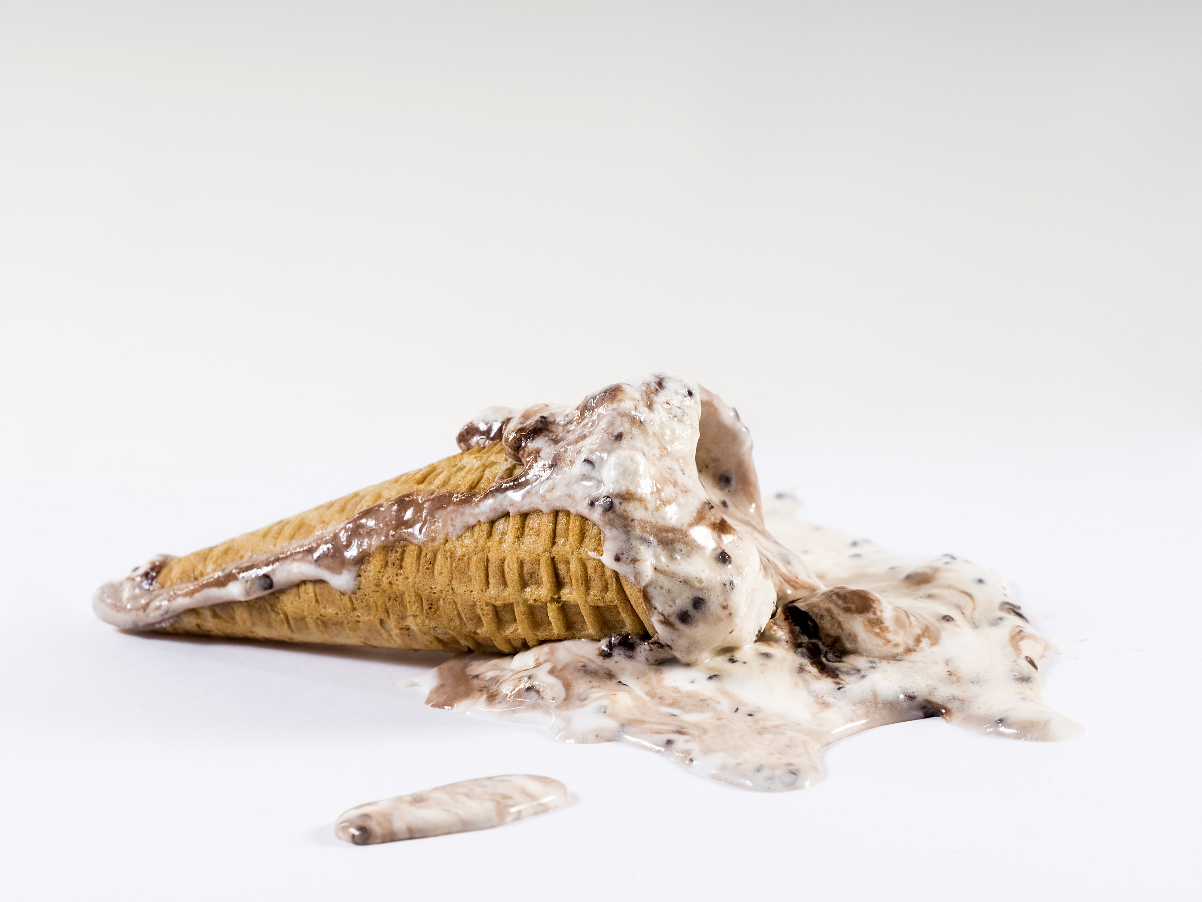 An ice cream cone tipped over with melted ice cream and chocolate pieces on a white surface