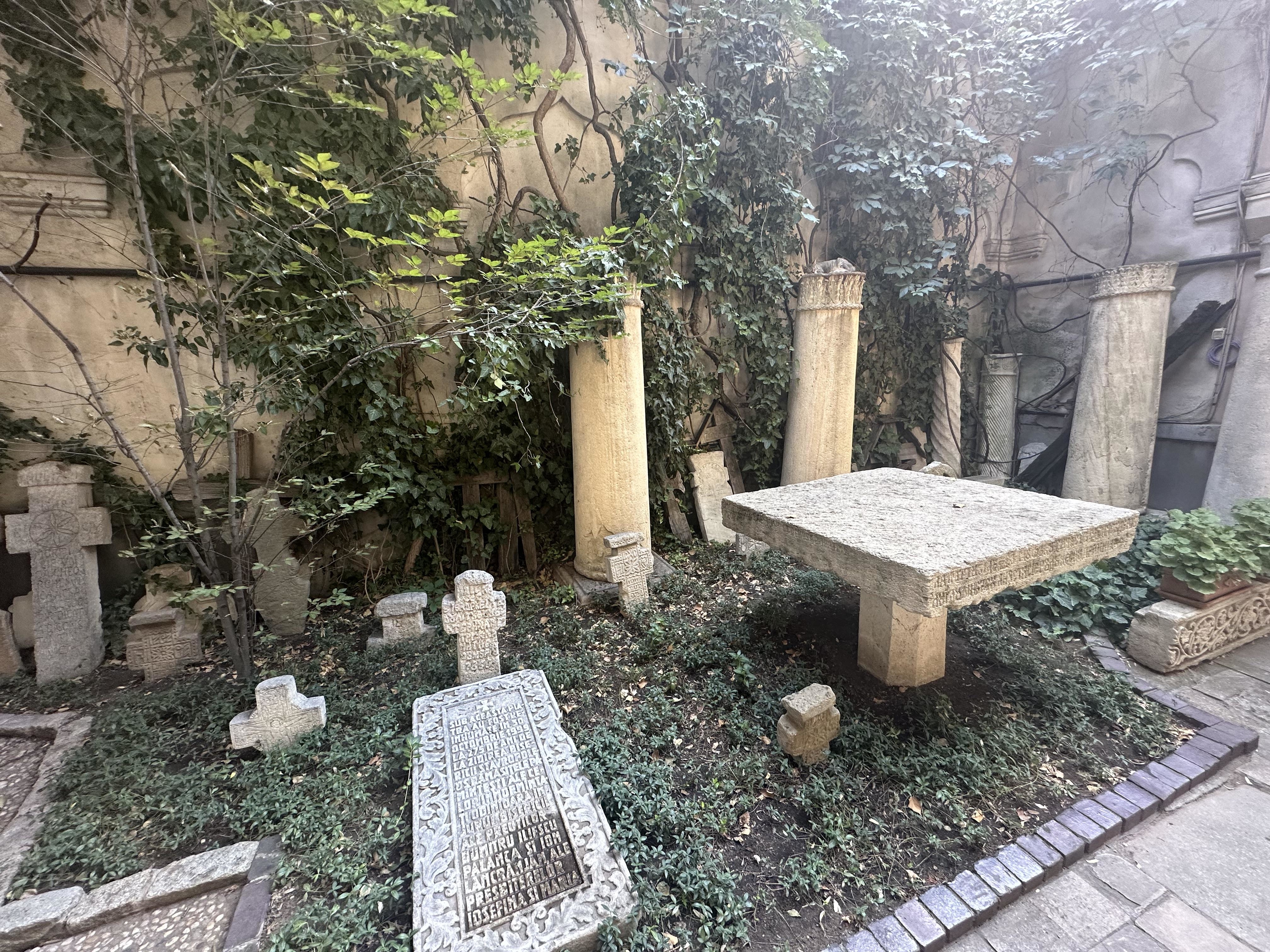 A small, aged cemetery with various gravestones and overgrown plants. A substantial tombstone table stands prominently in the center