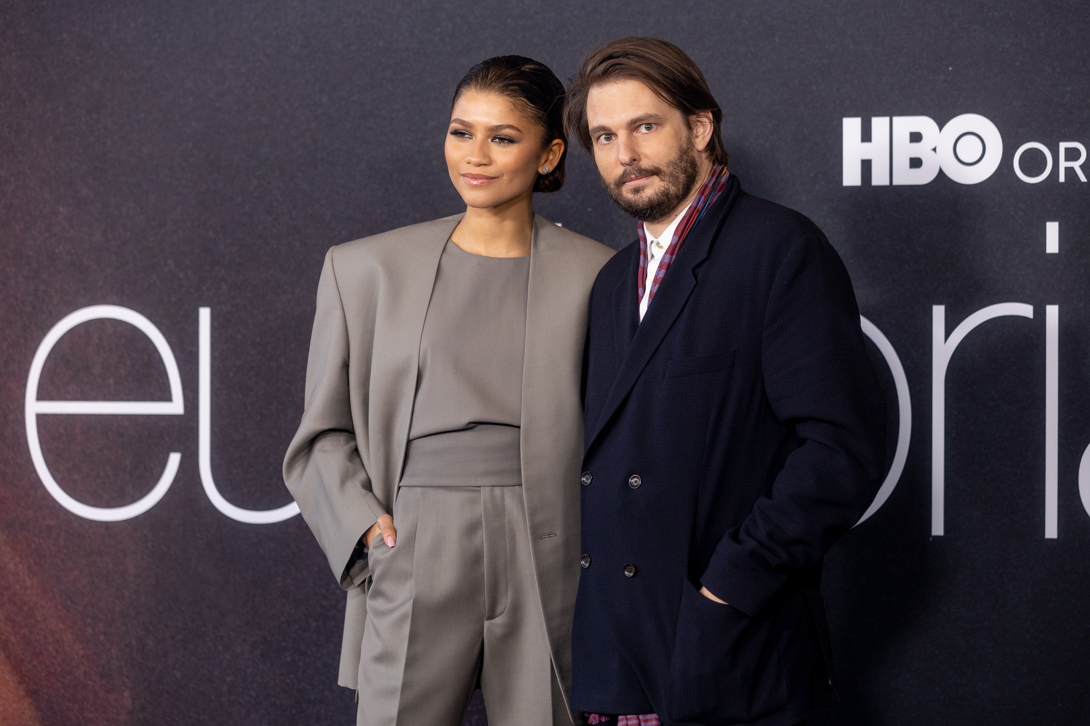 Zendaya in a chic pantsuit with hands in pockets, stands beside Sam Levinson, who is wearing a dark suit, at an HBO 'Euphoria' event red carpet
