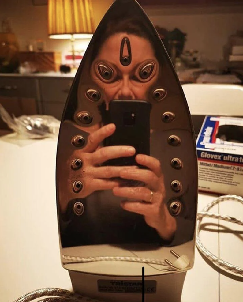 A woman takes a selfie with her phone reflected in the surface of an iron