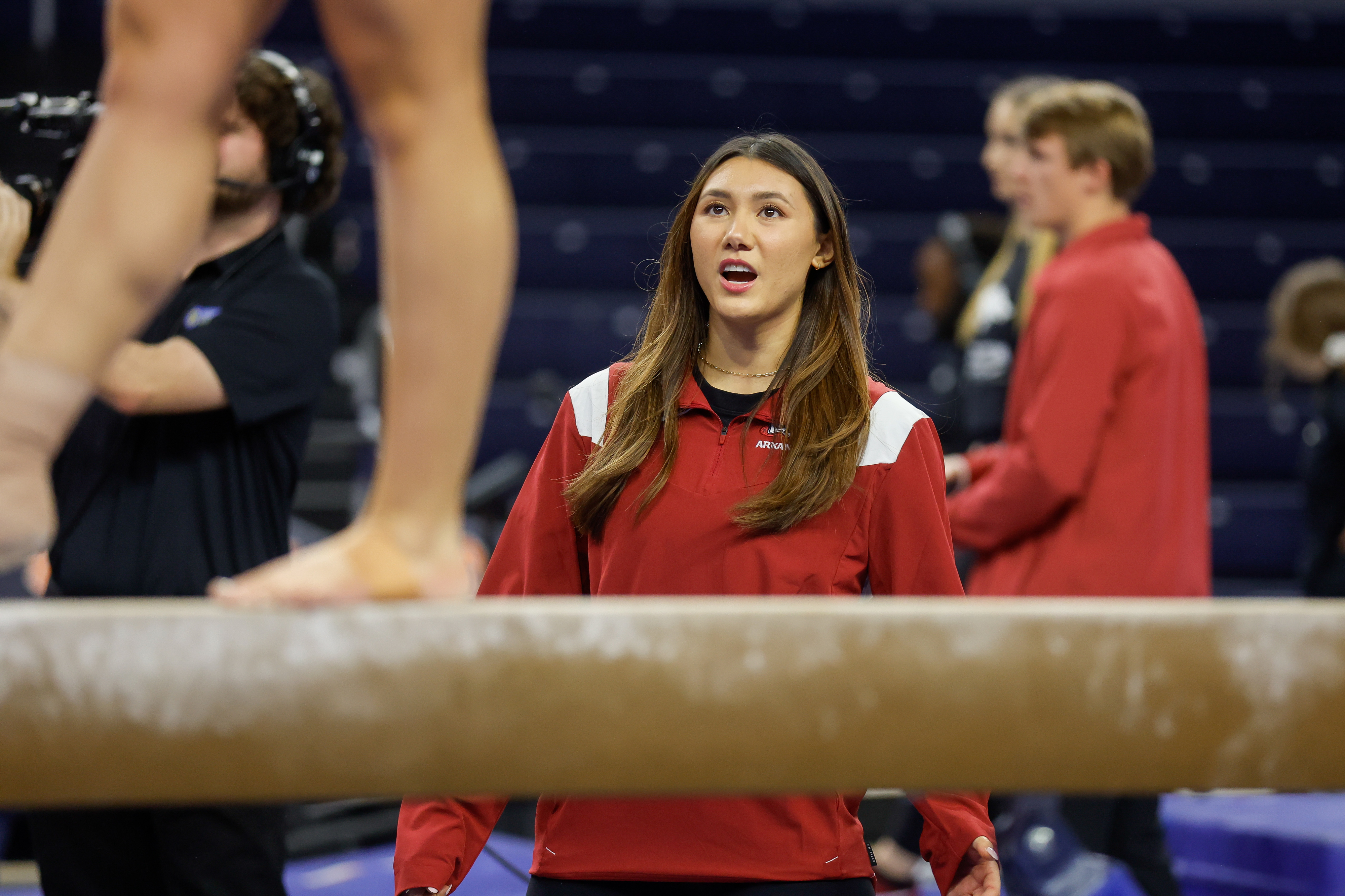 Kyla Ross watches an athlete on the balance beam during a gymnastics event, wearing a red and white jacket