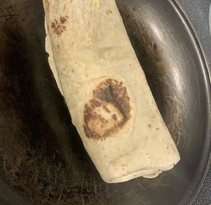 A burrito cooking in a pan with a face-like shape burnt into its surface