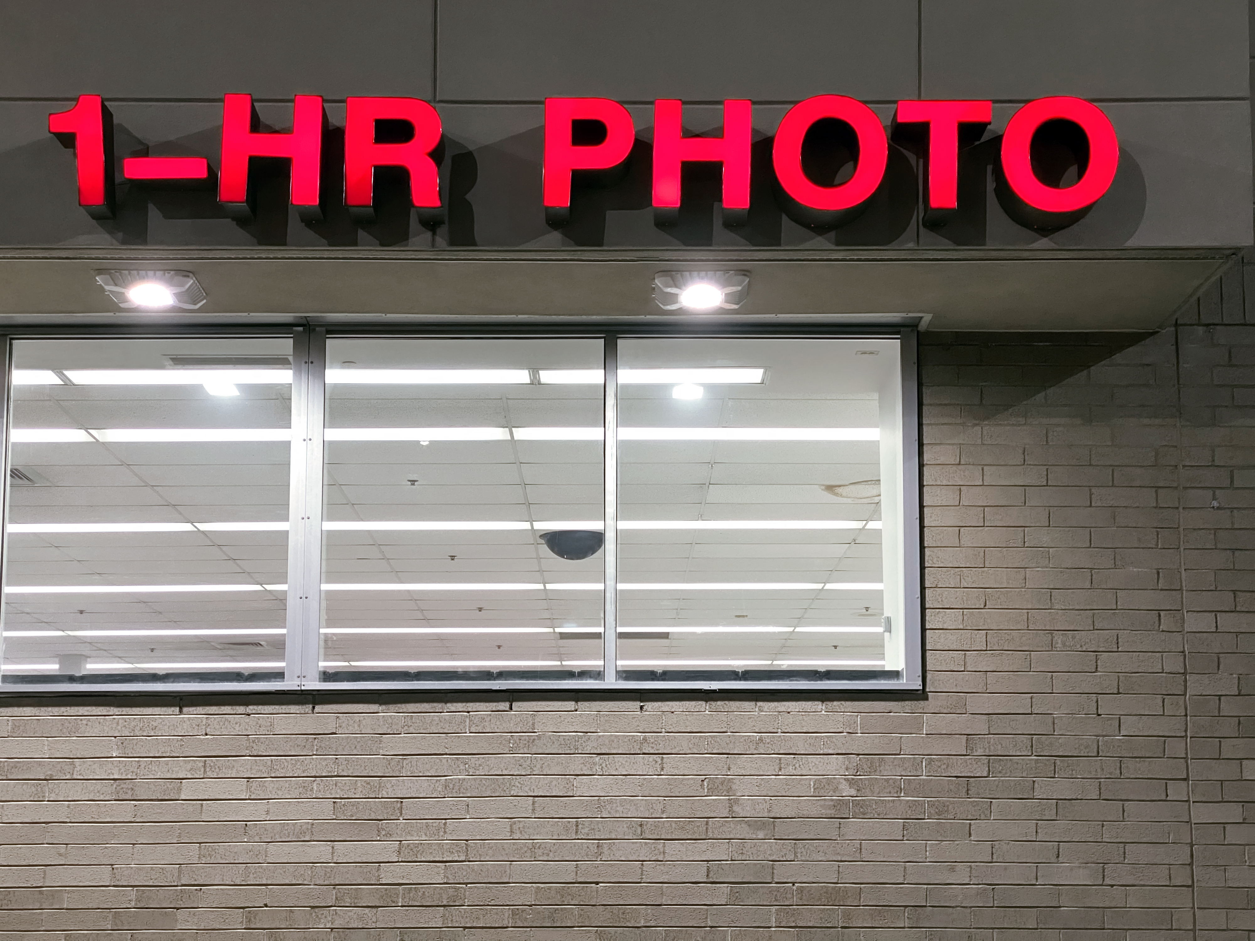 Exterior of a "1-HR PHOTO" store with a brick wall and lit windows showcasing the interior lights