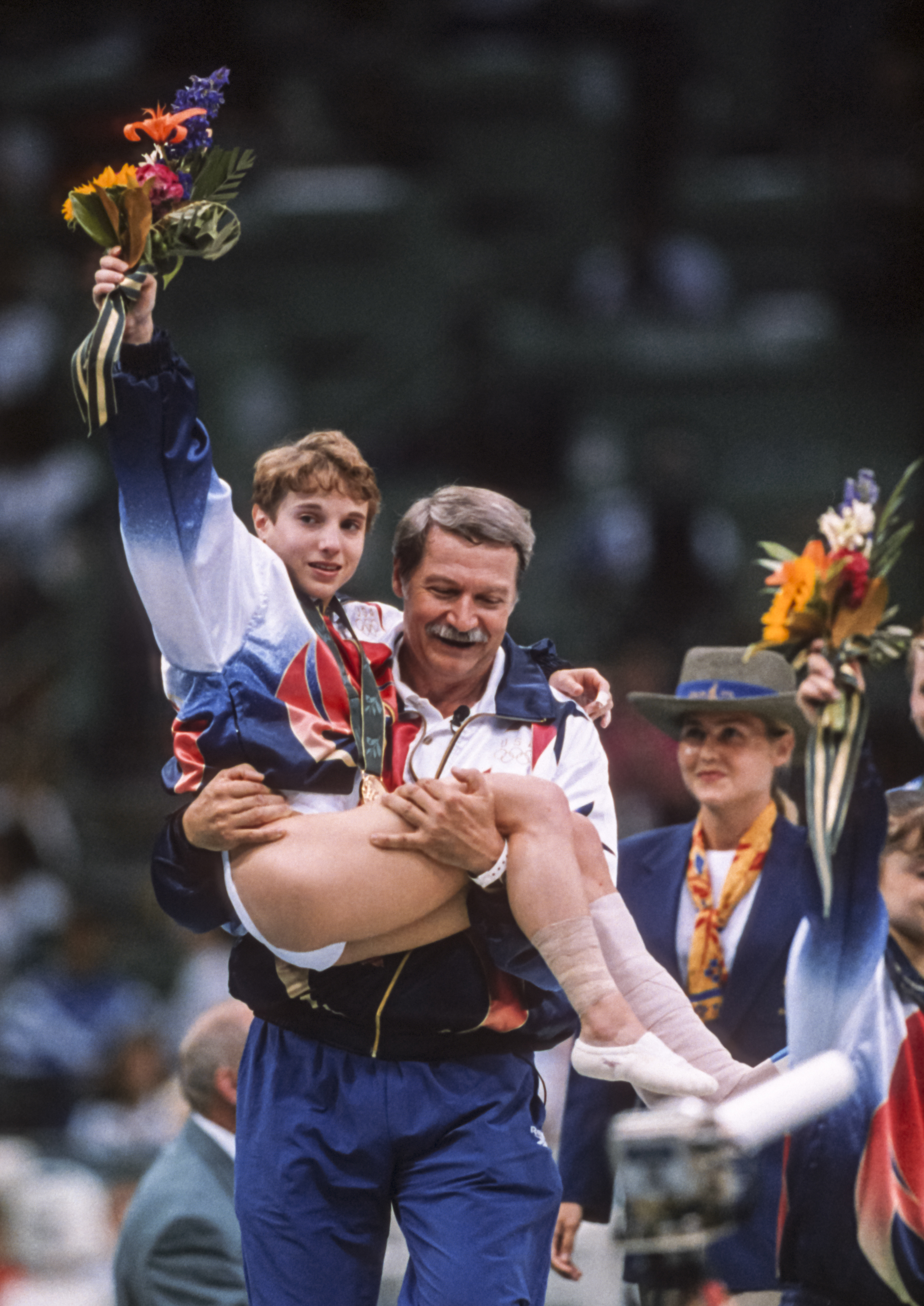 A young athlete holding flowers is carried by a man in a sports jacket at an event, with people celebrating in the background