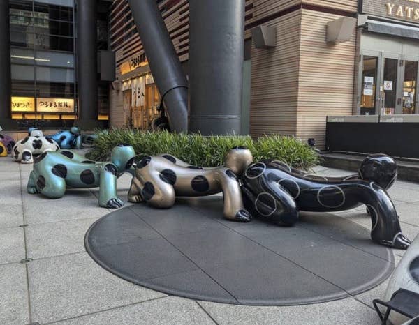 Sculptures of crawling humanoid figures with polka dots arranged in a line outside a building in an urban area
