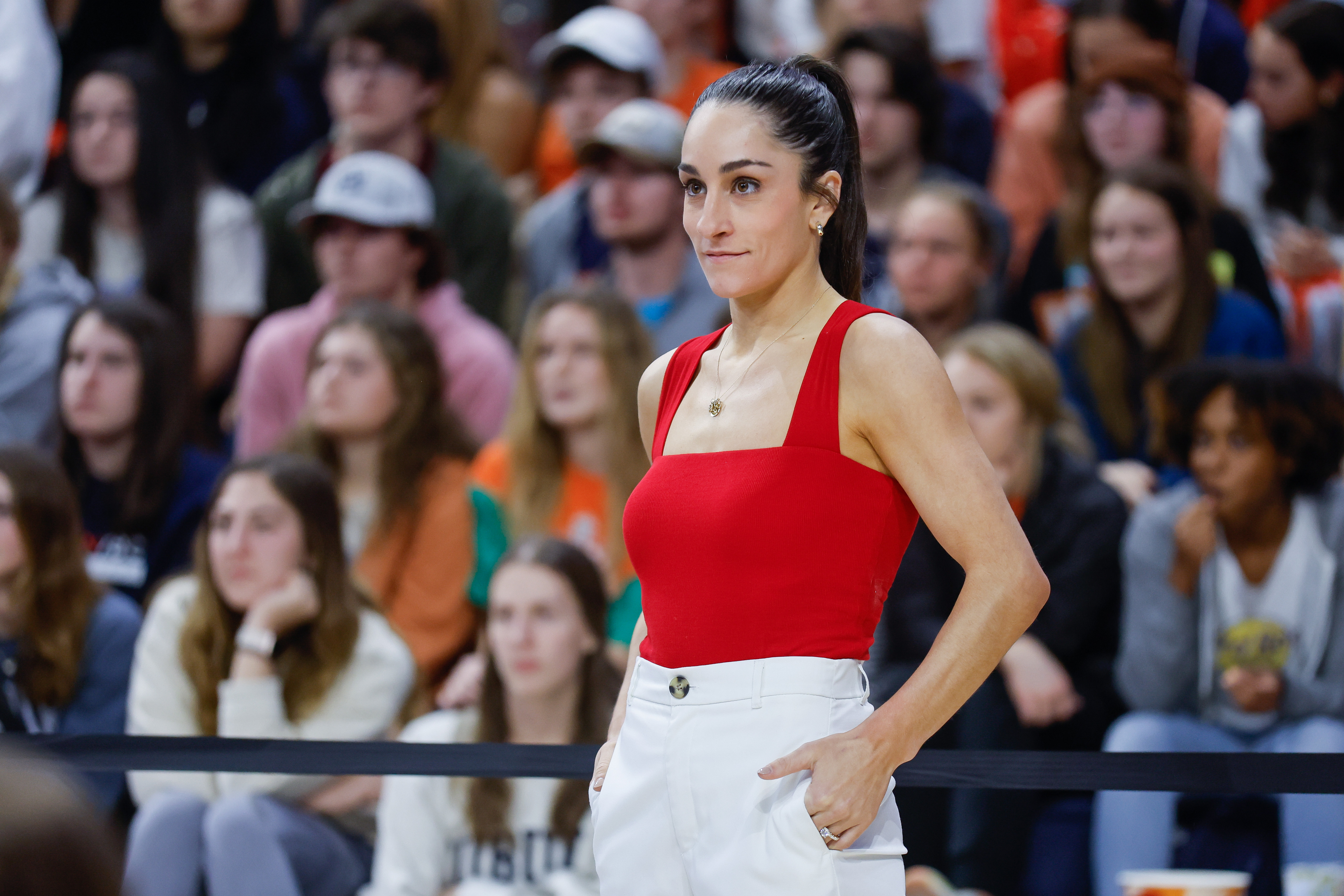 A woman in a red sleeveless top and white pants stands with her hands on her hips in front of a seated crowd
