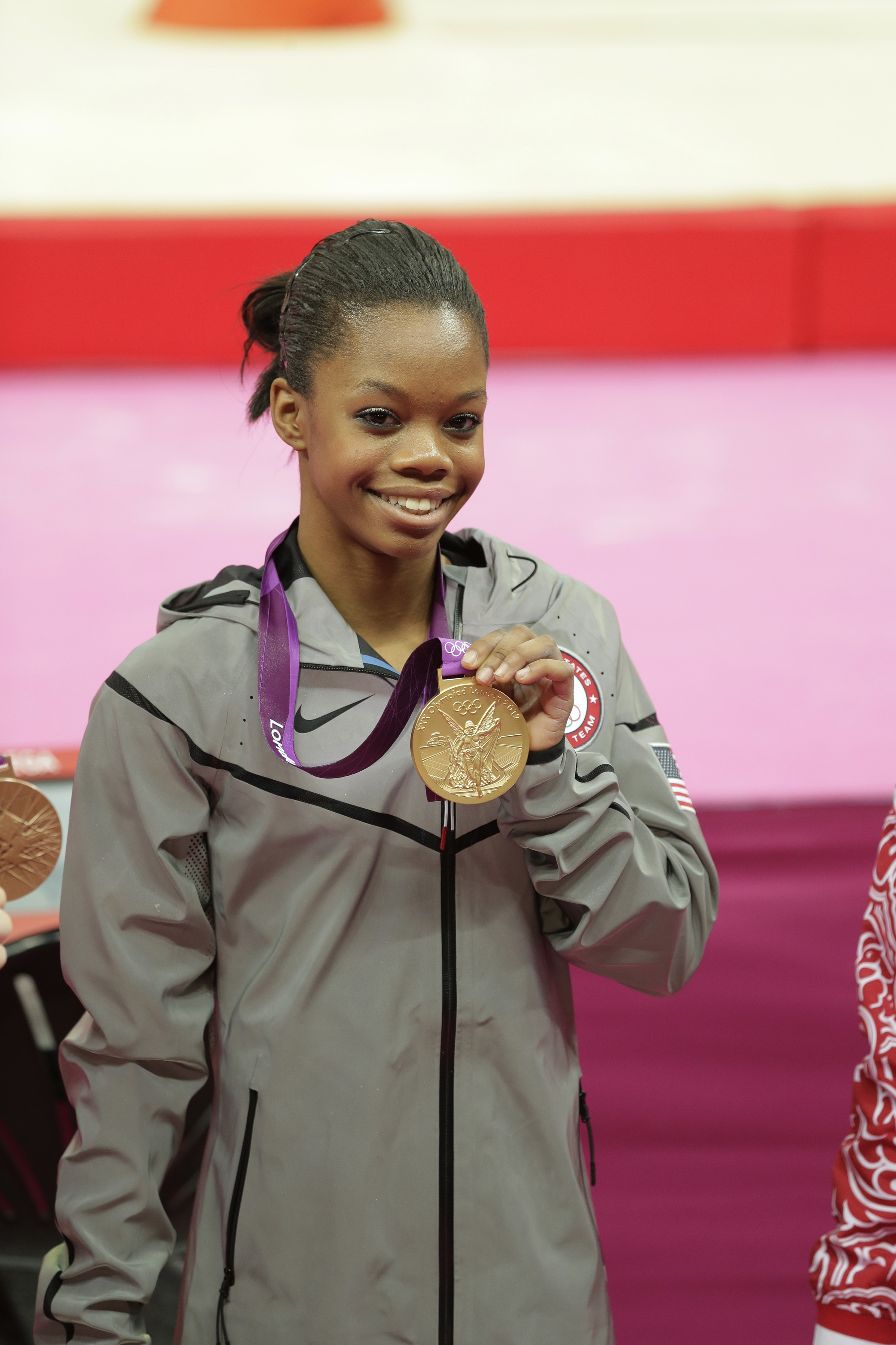 Gabby Douglas proudly holds up her Olympic medal while wearing a gray tracksuit
