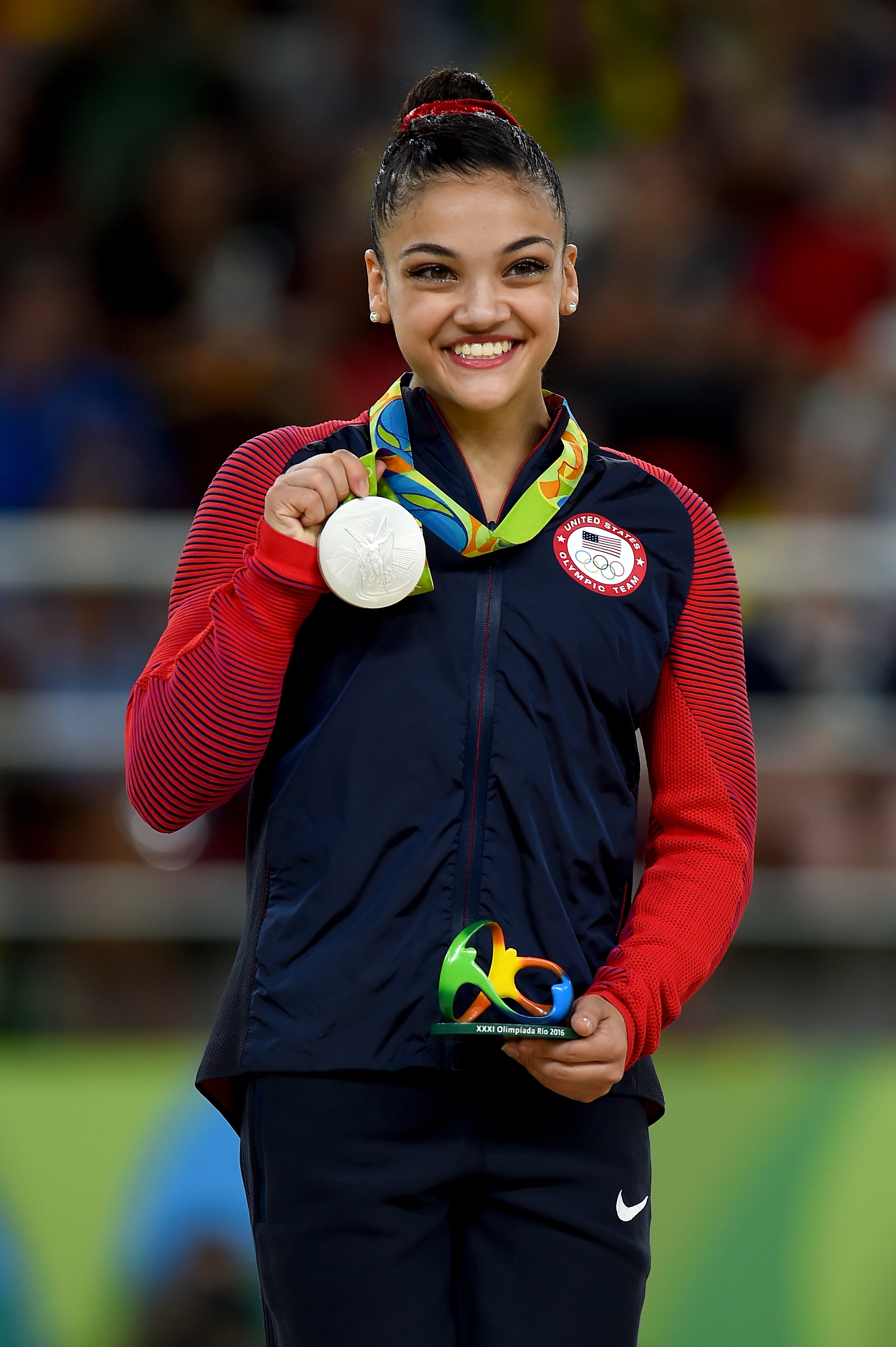 Laurie Hernandez smiles while holding a silver Olympic medal and a small trophy, wearing a Team USA jacket