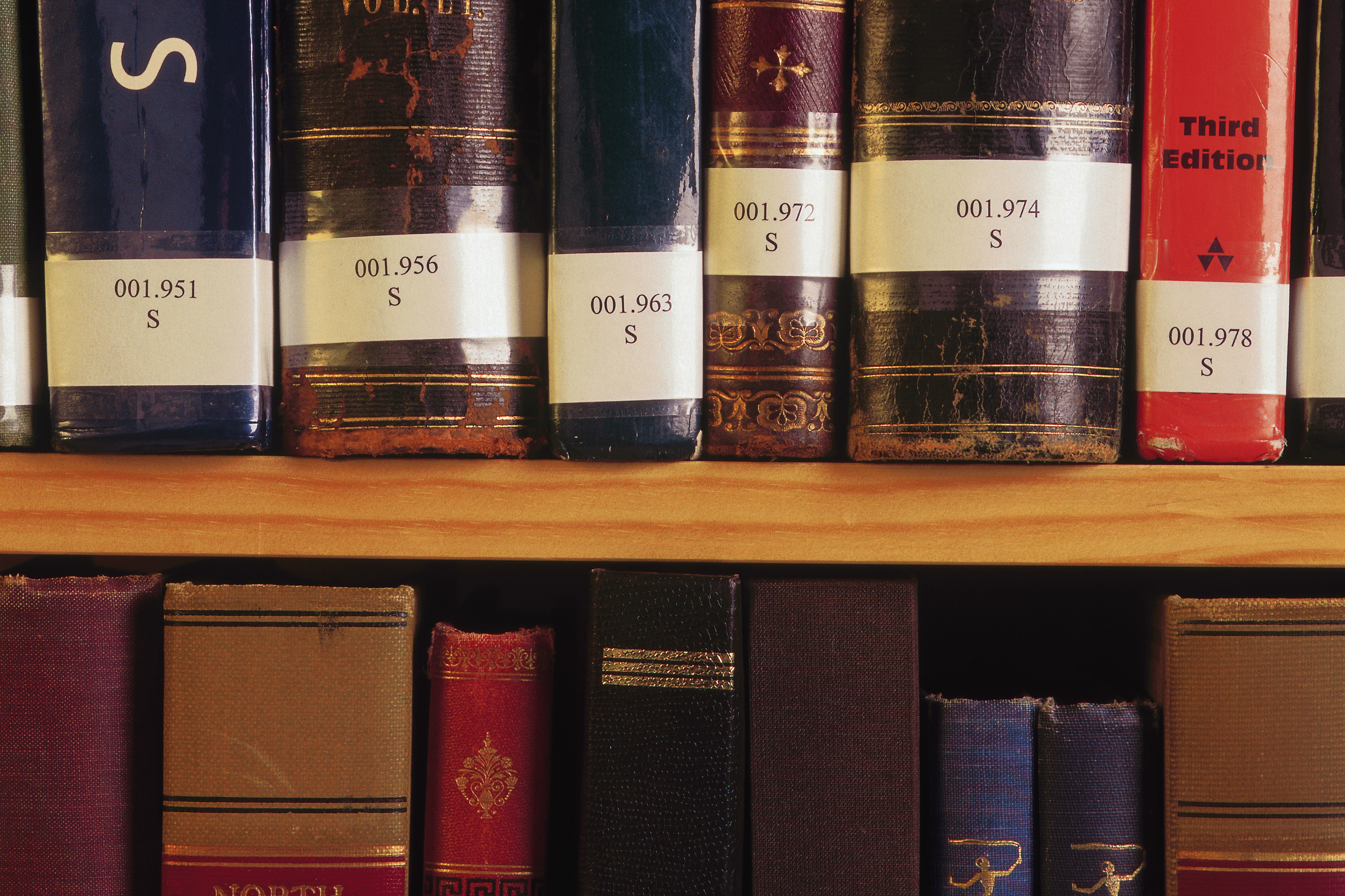 A close-up of two shelves of old, hardcover books, each with labels showing Dewey Decimal numbers and a letter "S" underneath. Some book titles are partially visible