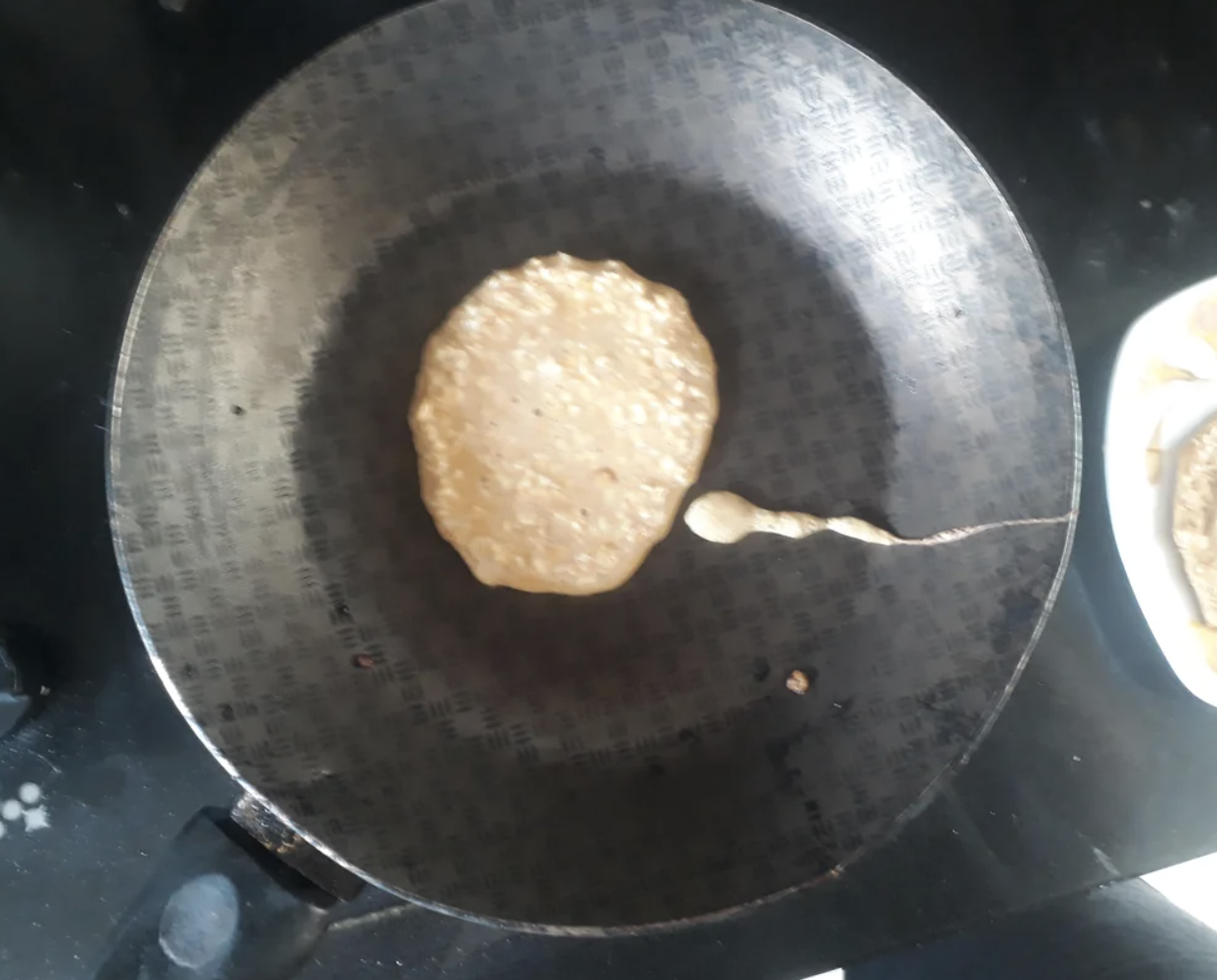 A pancake is being cooked in a frying pan on a stovetop. A small drop of batter is visible near the pancake