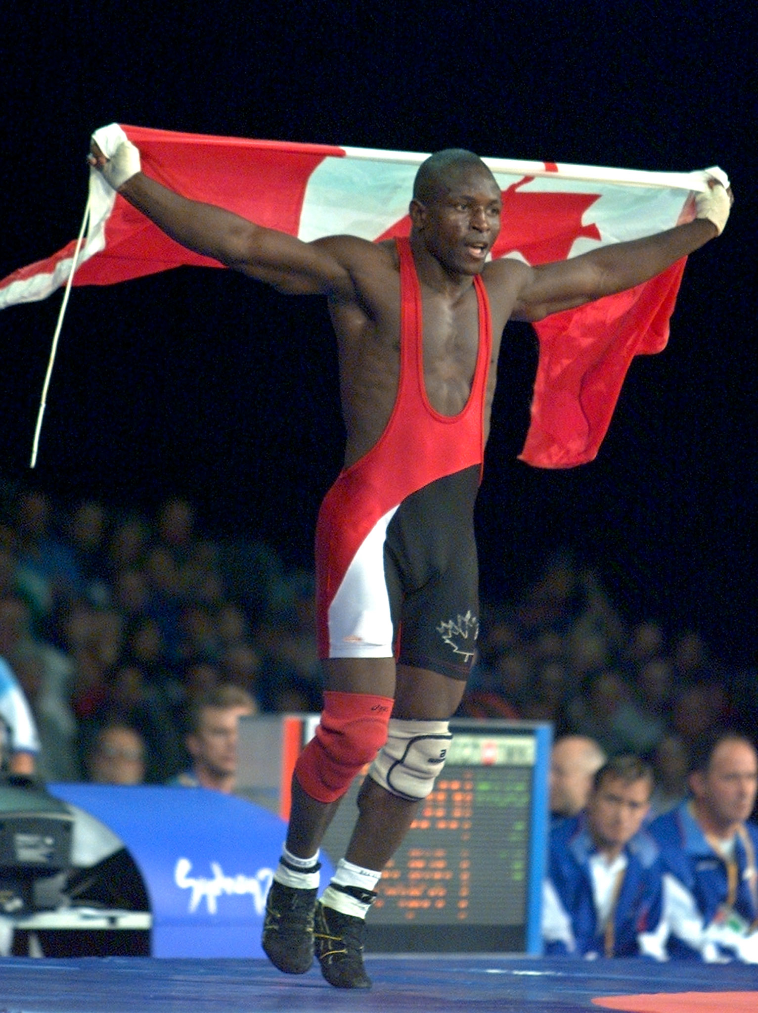 A wrestler holding a Canadian flag celebrates after a match. He is wearing a singlet and knee braces. There are people in the background watching