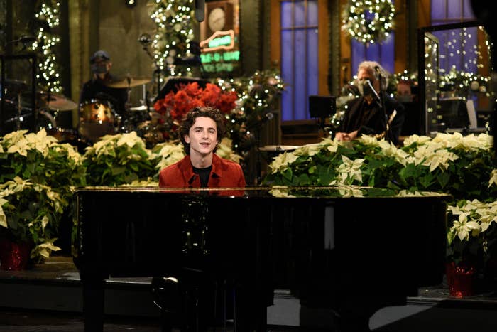 Timothée Chalamet plays piano, surrounded by holiday decor, during a performance on a stage. Musicians are visible in the background