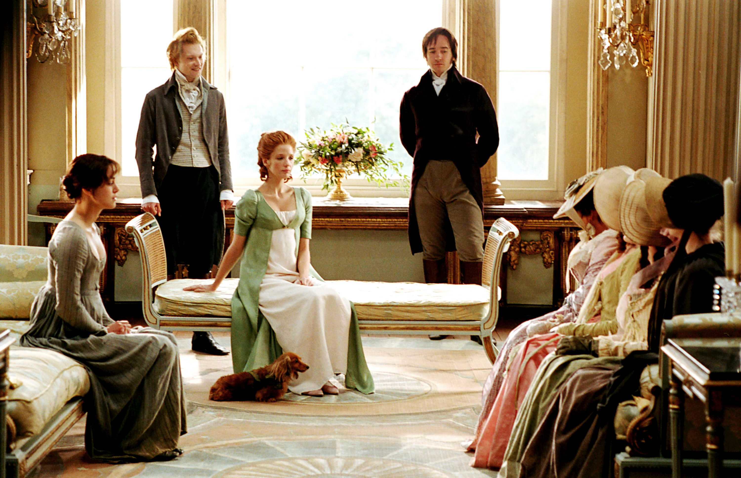 Cast of "Pride and Prejudice" in period costumes. The women wear elegant dresses and the men are in 19th-century attire, seated in a luxurious room
