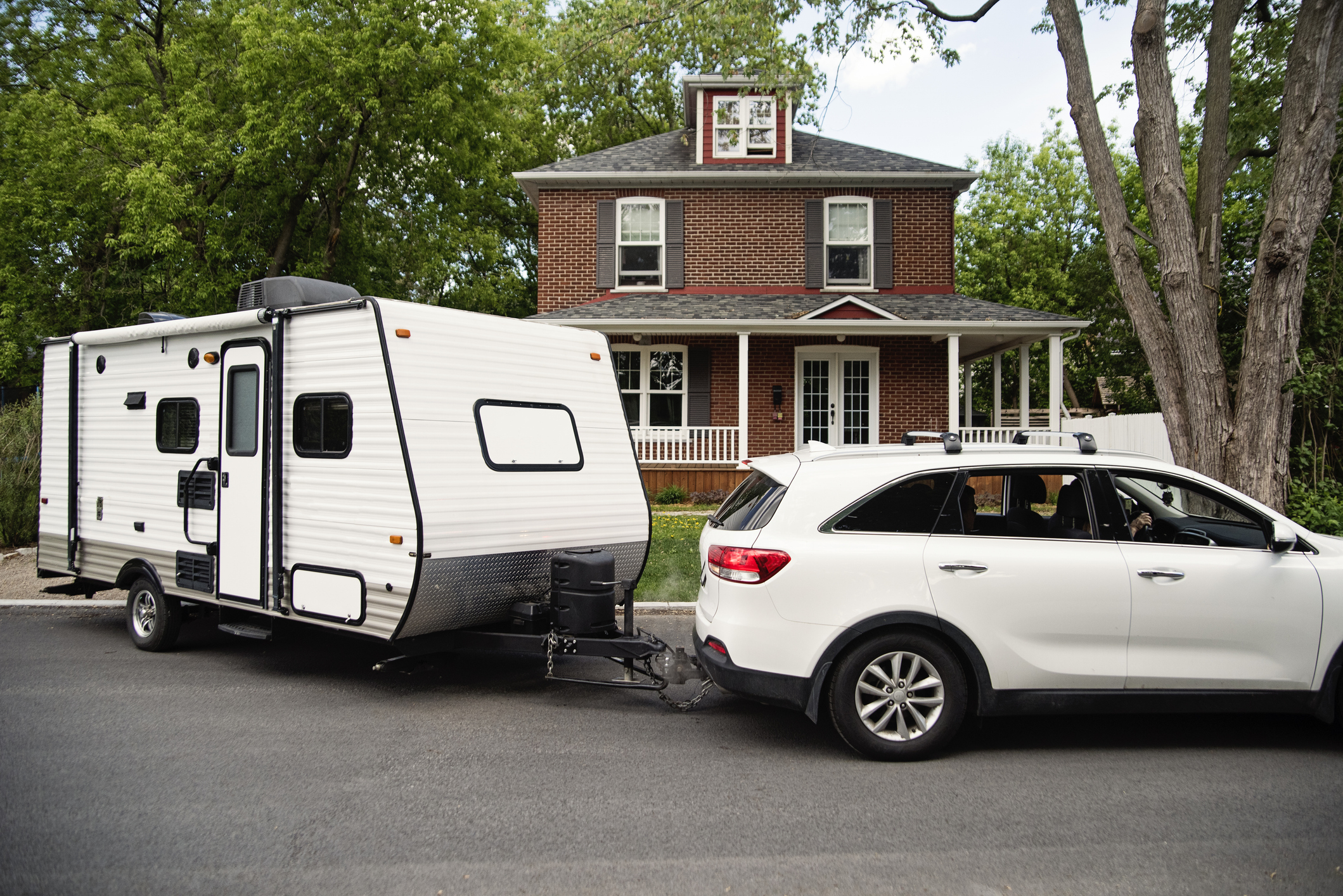 A white SUV is towing a small travel trailer in front of a two-story brick house
