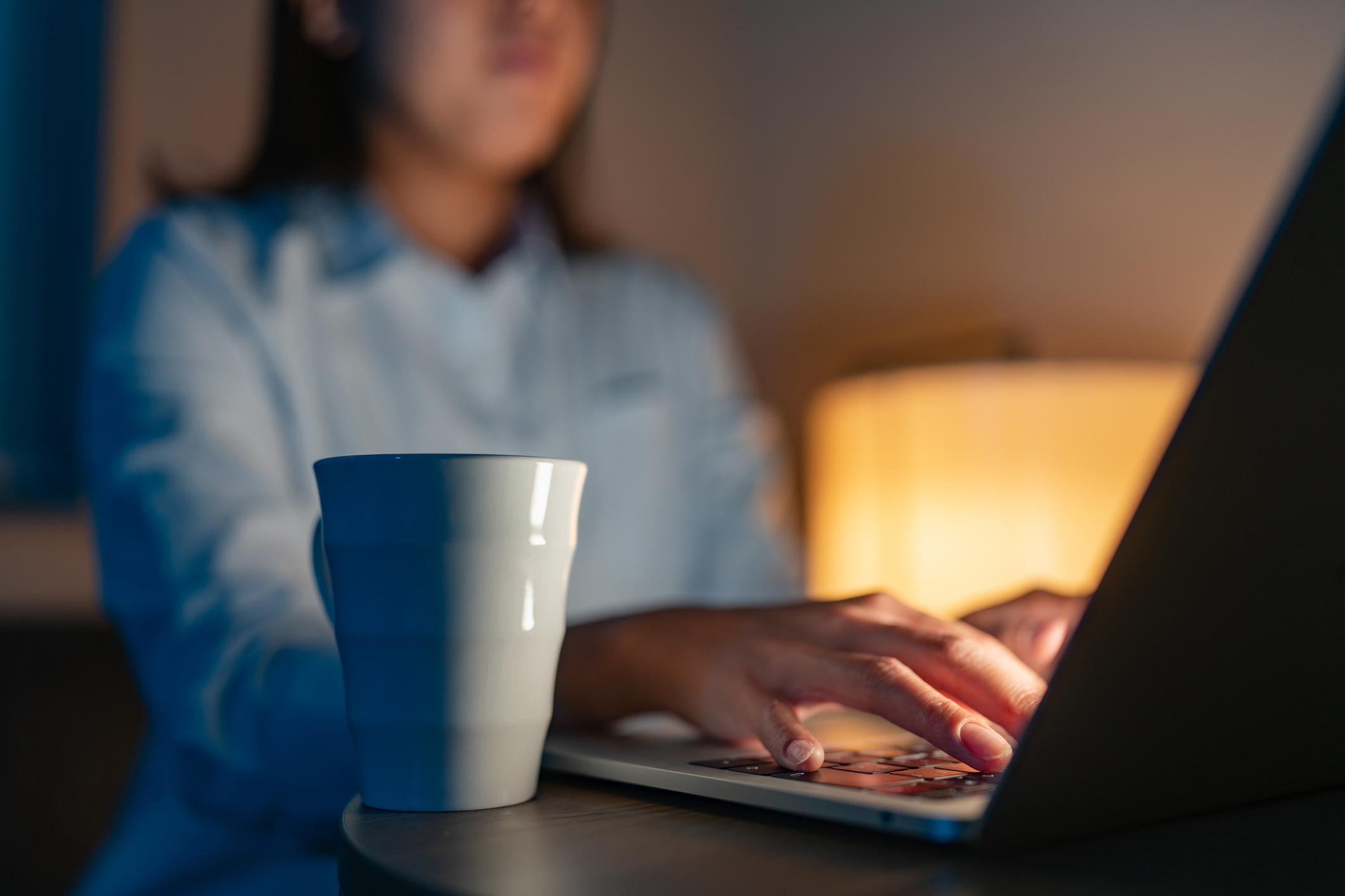 Person typing on a laptop with a mug placed on a nearby table. Background includes a softly lit lamp. The image is in a calm, indoor setting