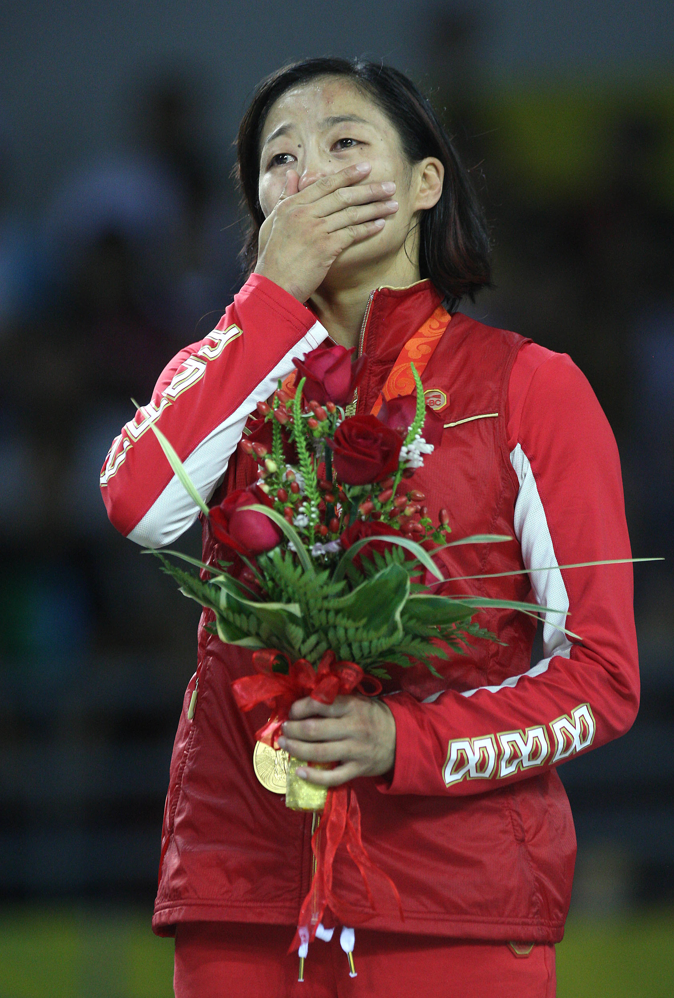 Female athlete in a red tracksuit with &quot;China&quot; on the sleeve, holding flowers and a gold medal, hand over mouth in an emotional moment