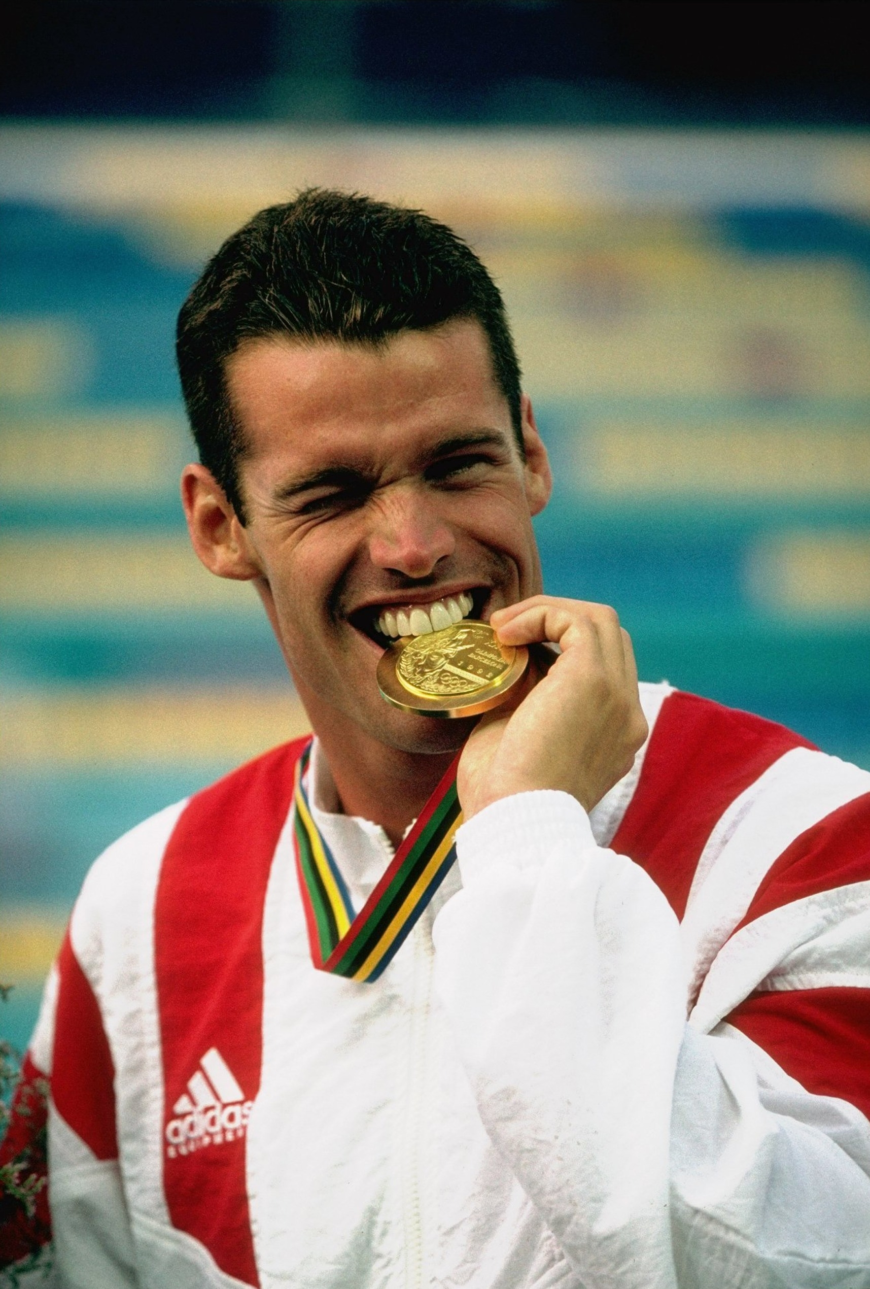 An athlete wearing a red and white Adidas jacket bites his gold medal, showing a joyful expression. Background appears to be a sports venue