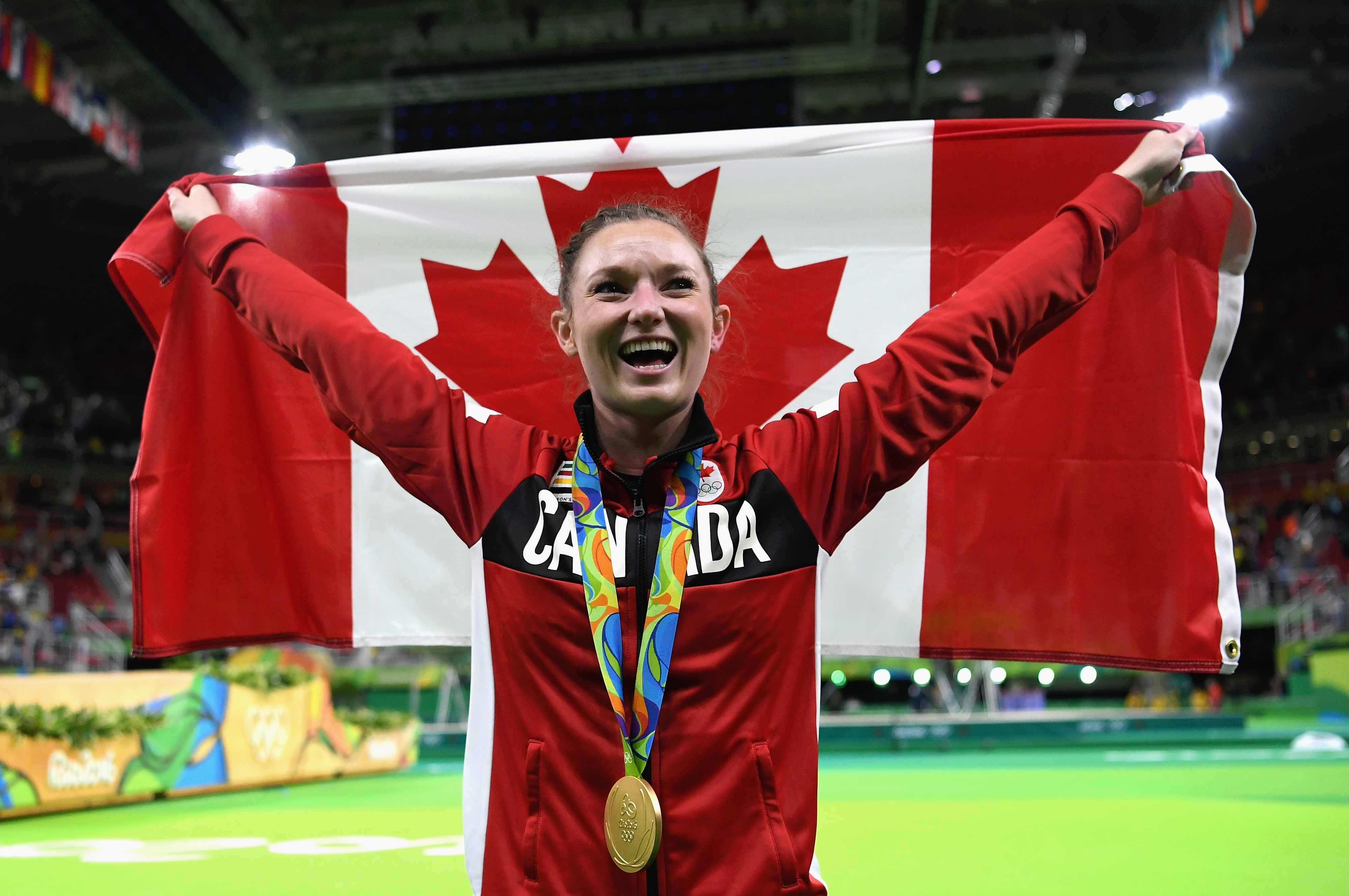 Canadian athlete stands jubilantly with a large Canadian flag draped behind, wearing a red jacket, displaying her gold medal at a sports event