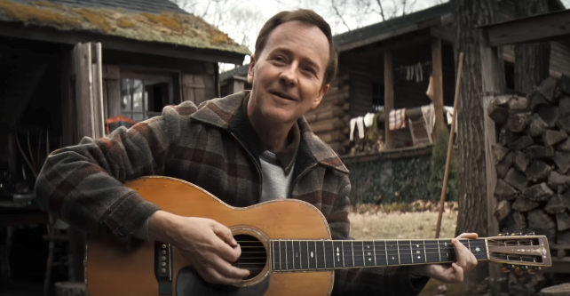 A man in a plaid jacket is sitting outside next to a pile of firewood, playing an acoustic guitar and smiling at the camera
