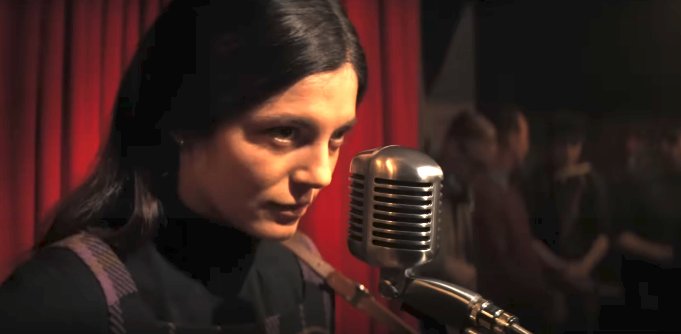 Unidentified woman speaks into a vintage microphone in front of a curtain. People stand blurred in the background