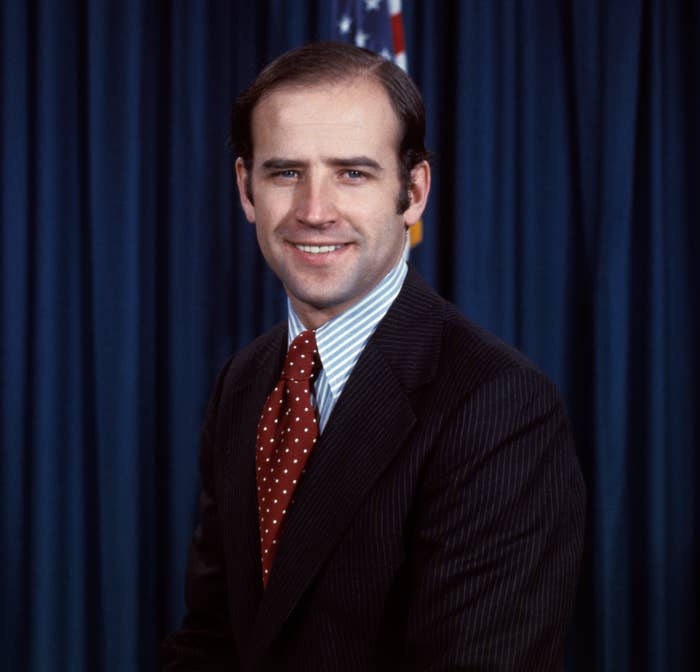A man in a suit with a polka dot tie stands in front of a curtain and an American flag, smiling