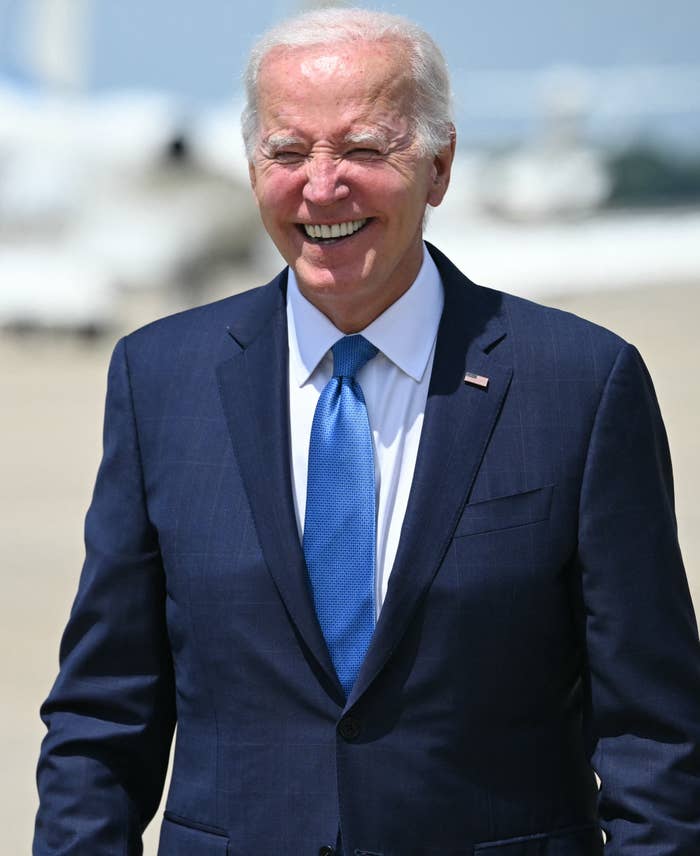 Joe Biden smiling and walking outdoors in a suit and blue tie. Airplane in the background