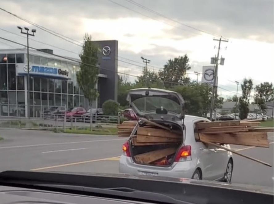 A small car with its trunk open and filled with long wooden planks that extend out of the vehicle, driving near a Mazda dealership