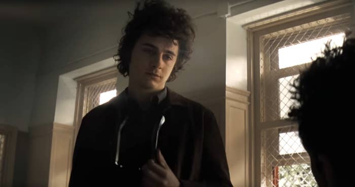 Timothée Chalamet, with tousled hair, stands indoors holding a stethoscope around his neck in a scene from a TV show or movie