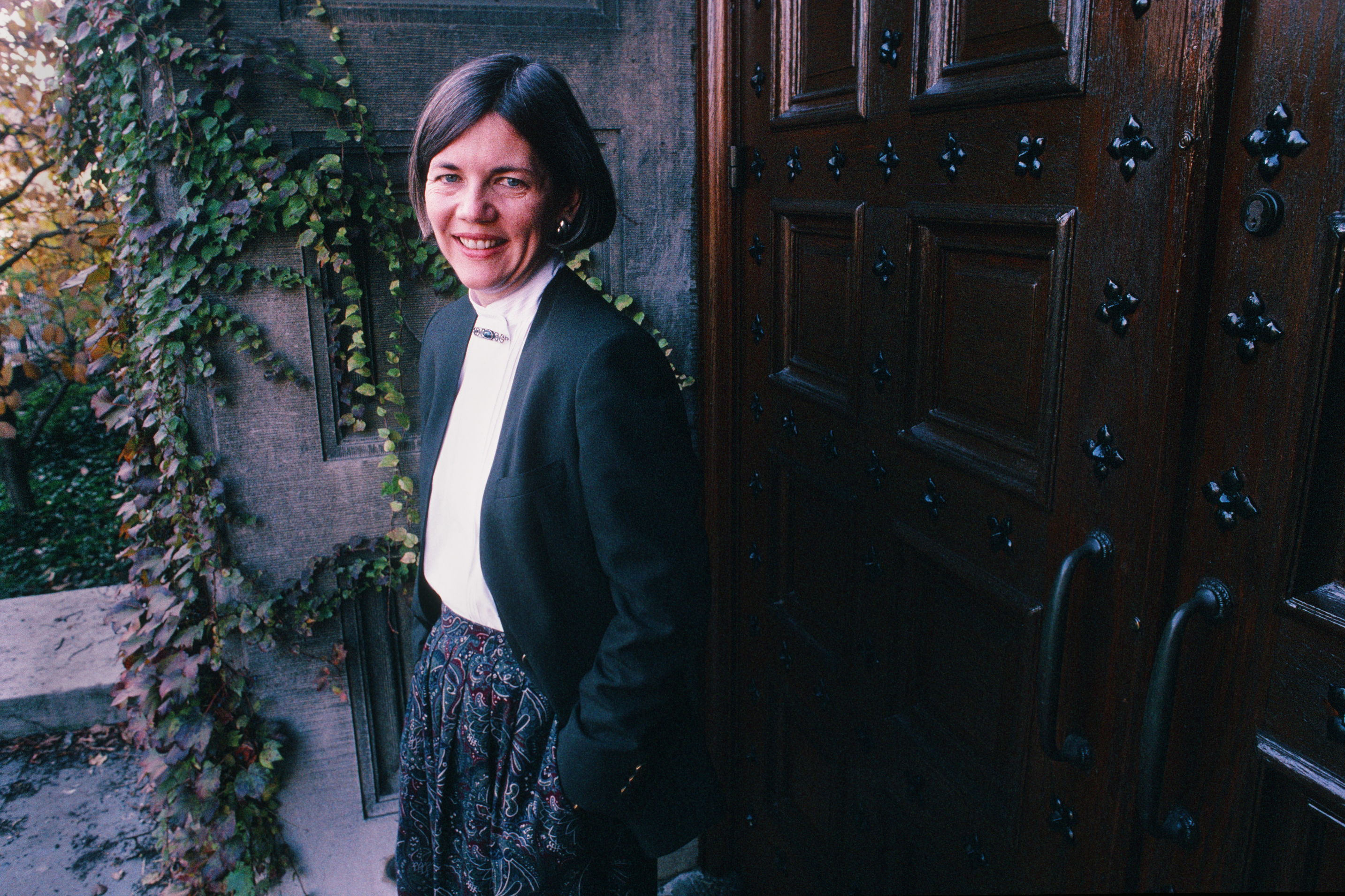 A woman standing outdoors next to a wooden door covered in decorative ironwork, wearing a blazer and a patterned skirt, smiling at the camera. Name unknown