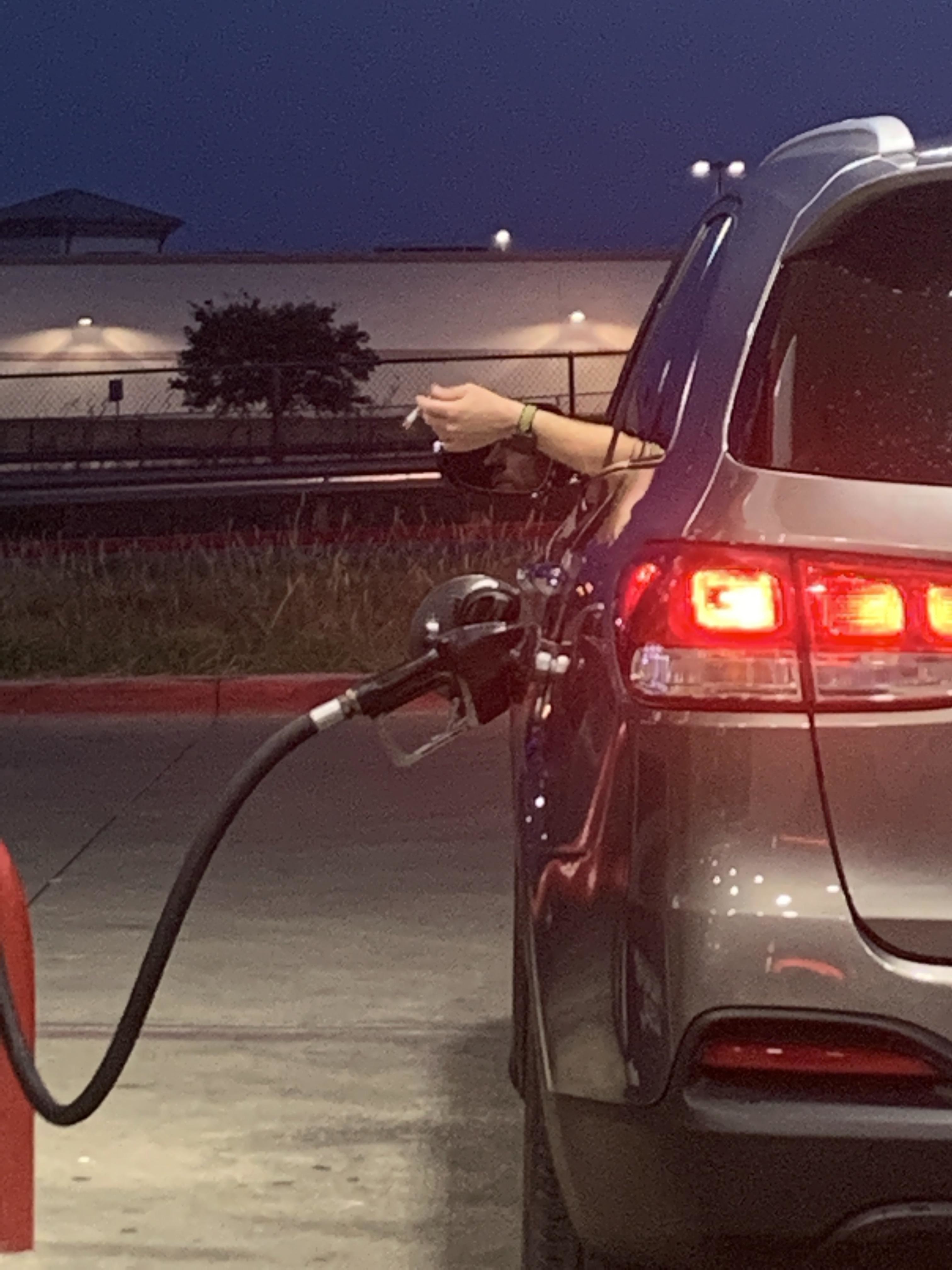 A person smokes a cigarette while sitting in a vehicle at a gas station, with the fuel nozzle still inserted in the car’s gas tank