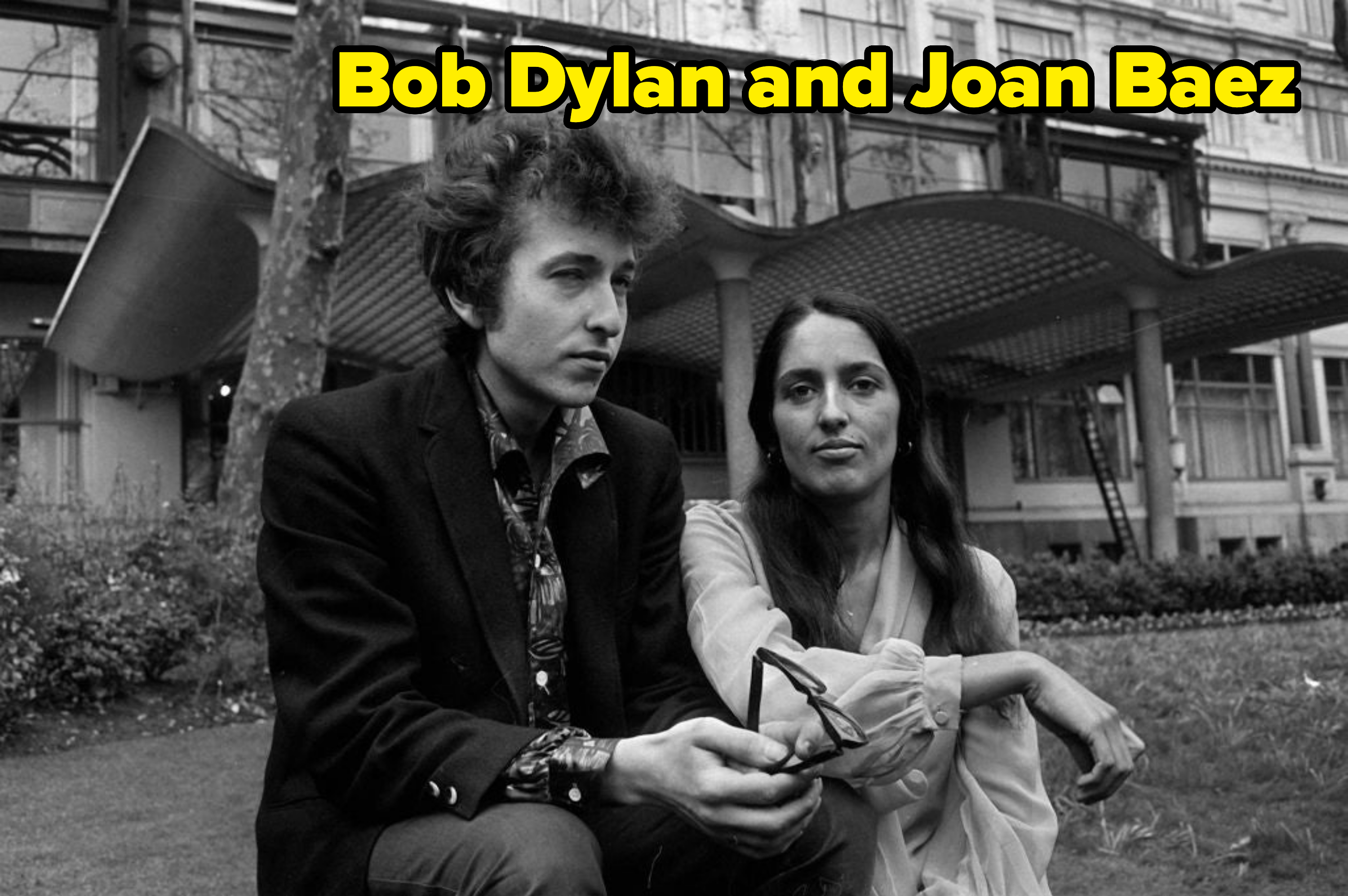 Bob Dylan and Joan Baez sitting together on grass in front of a building, with Dylan holding sunglasses