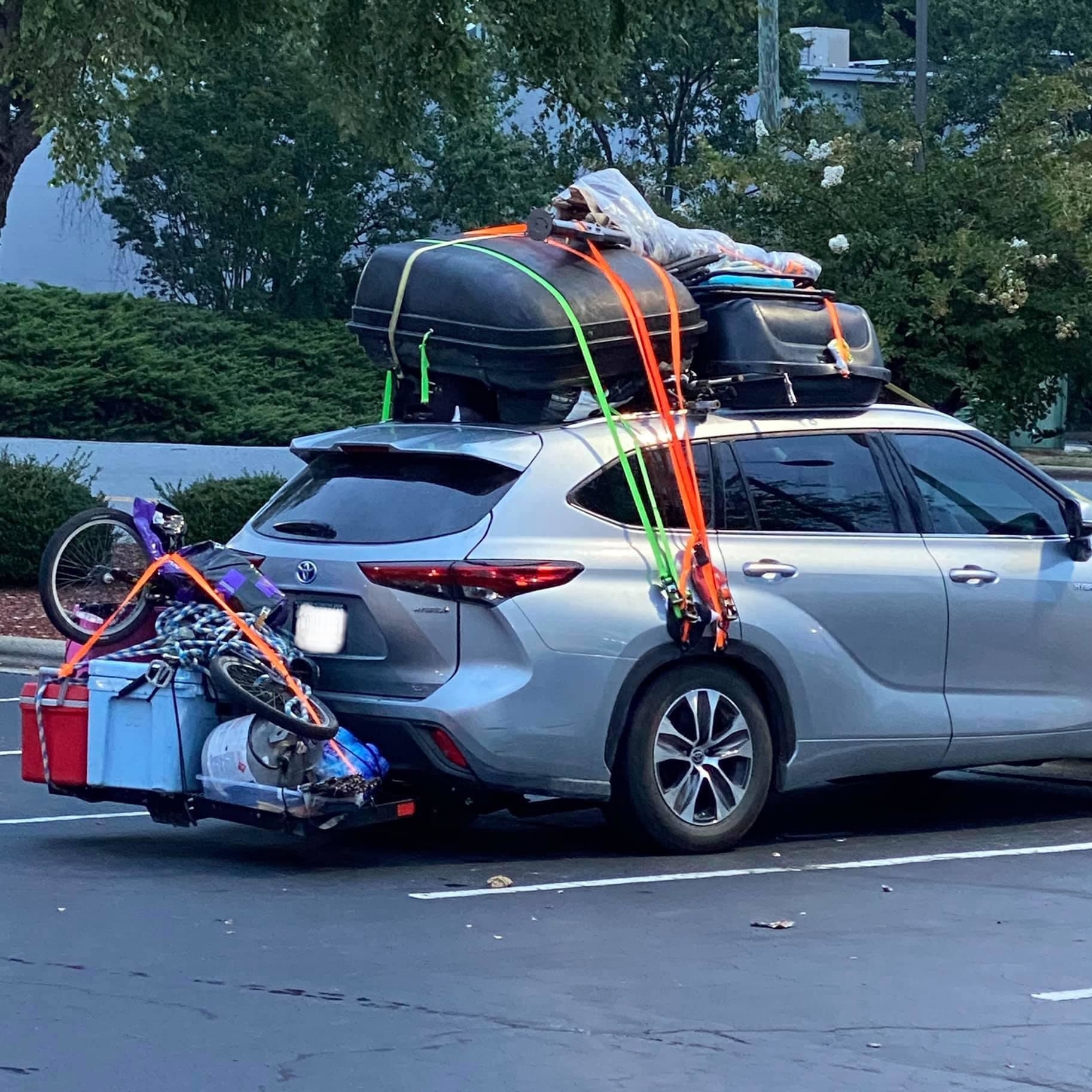 Silver SUV packed for travel with items including a bike, cooler, and boxes strapped to the rear and roof, secured with bright orange straps in a parking lot