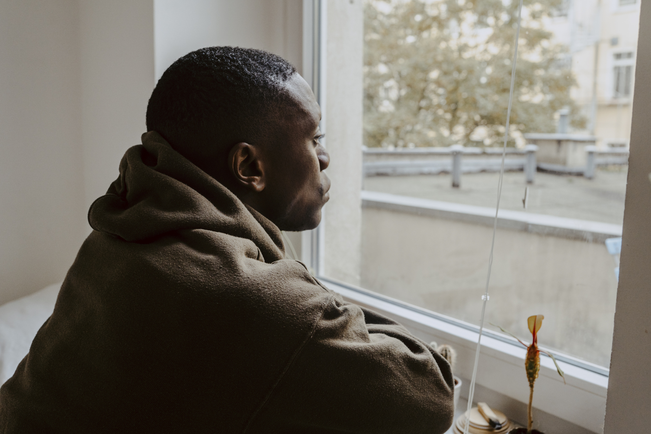 Man in a hoodie gazes out a window, lost in thought. The scene evokes introspection and solitude. Background shows an urban environment