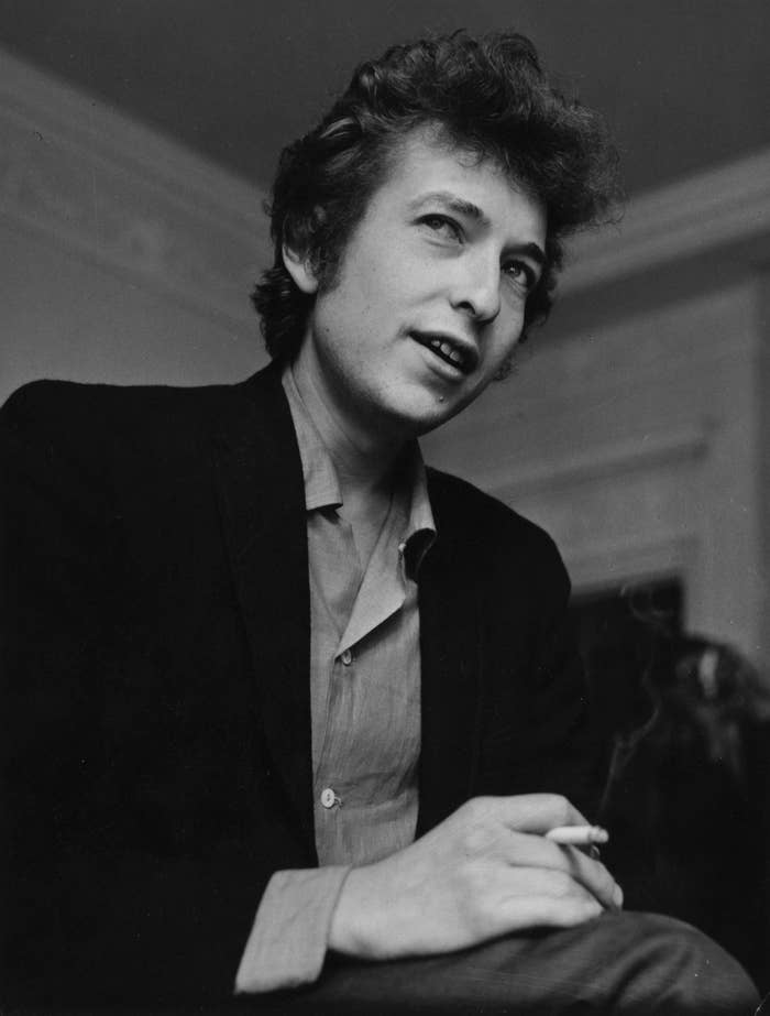 Bob Dylan holding a cigarette, dressed in a casual shirt and jacket