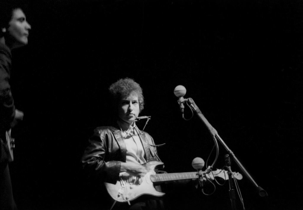Bob Dylan on stage holding an electric guitar, wearing a leather jacket, with a microphone stand in front of him. Another unidentified person is partially visible