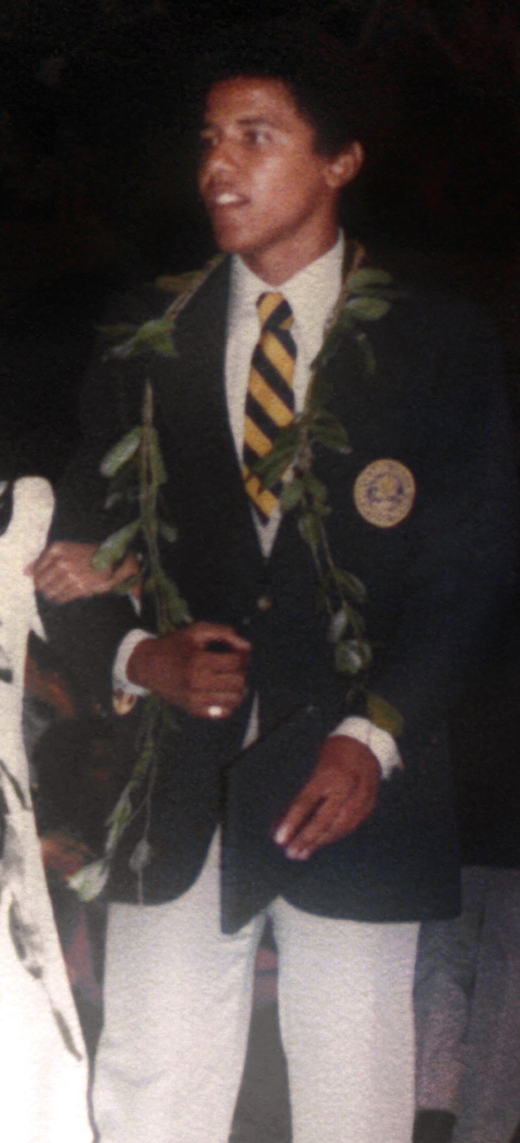 A young woman in a long white dress and floral garland smiles as she walks beside a young man in a dark blazer and tie, also wearing a garland