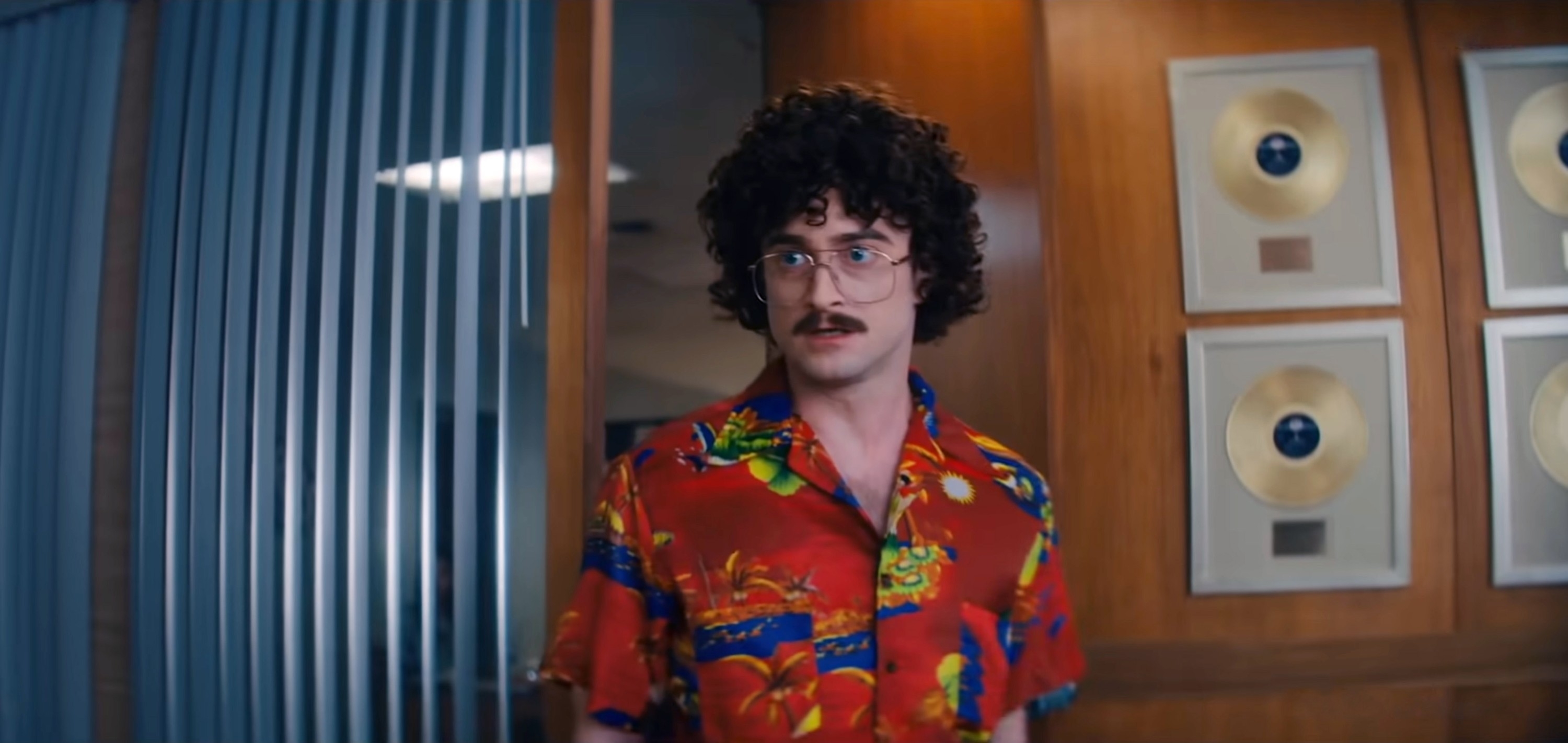 Daniel Radcliffe dressed as Weird Al Yankovic, wearing a patterned shirt, standing in front of a wall with gold records