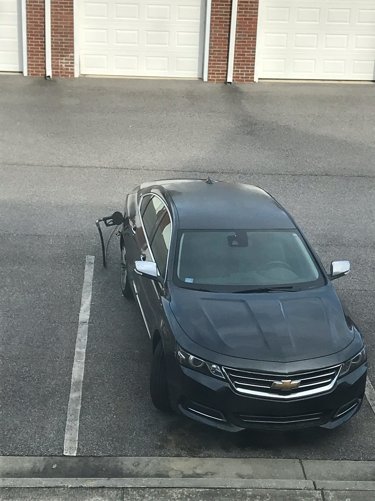 A gray Chevrolet sedan is shown parked with a fuel pump nozzle hanging from its gas tank, probably forgotten