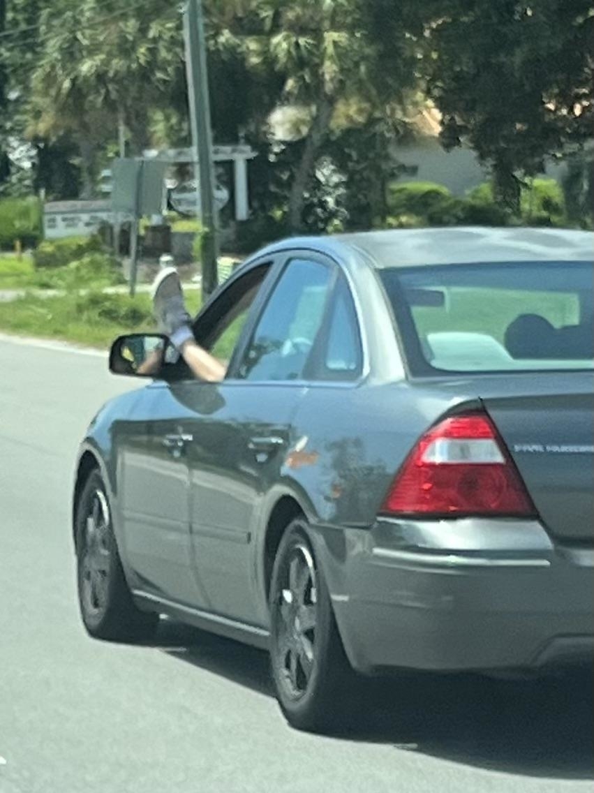 A person in a car extends their foot through the driver's side window while driving on a road. Trees and street signs are visible in the background
